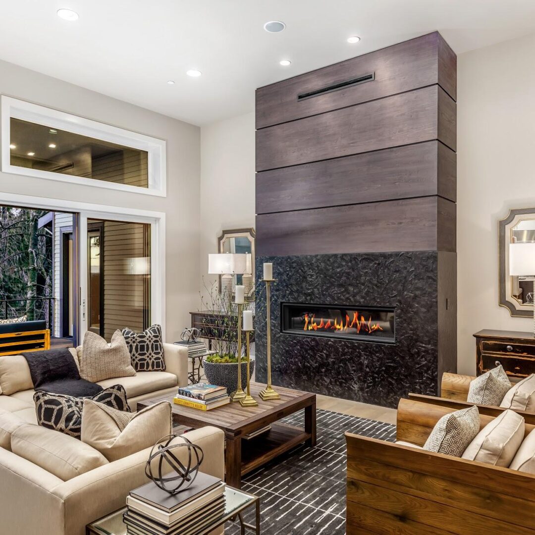 Modern living room with beige sofas, patterned pillows, wooden armchairs, a large coffee table, and a sleek fireplace with dark panels. Large windows and glass doors provide natural light and outdoor views.