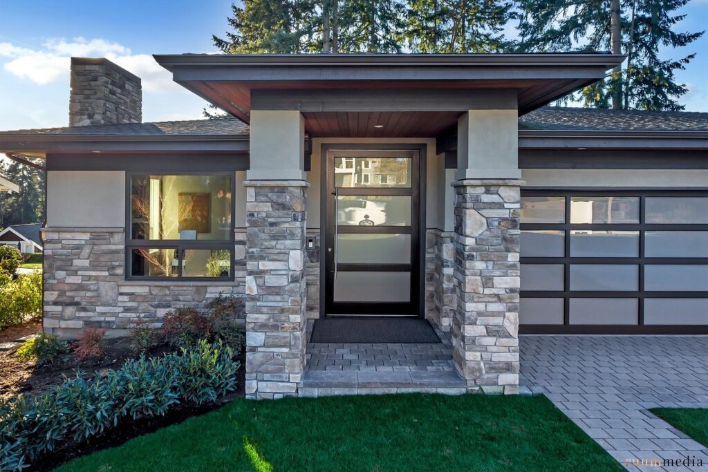 Modern house exterior with stone and wood accents, large glass windows, a glass front door, and a paved walkway surrounded by landscaped greenery. Tall trees are visible in the background.