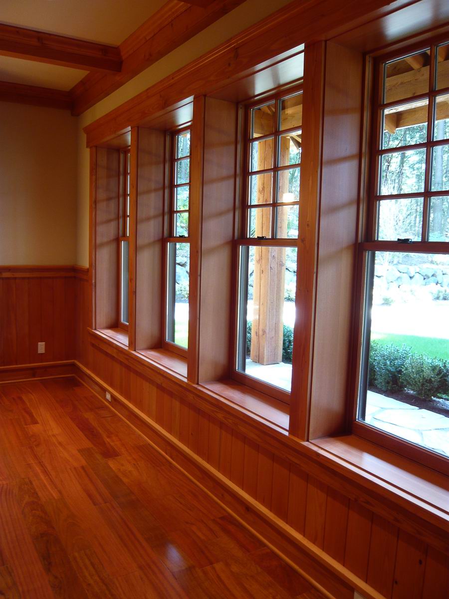 A row of large wooden-framed windows lets natural light into a room with polished wood floors, wood-paneled lower walls, and exposed wooden ceiling beams. A view of greenery and a wooden porch structure is visible outside.