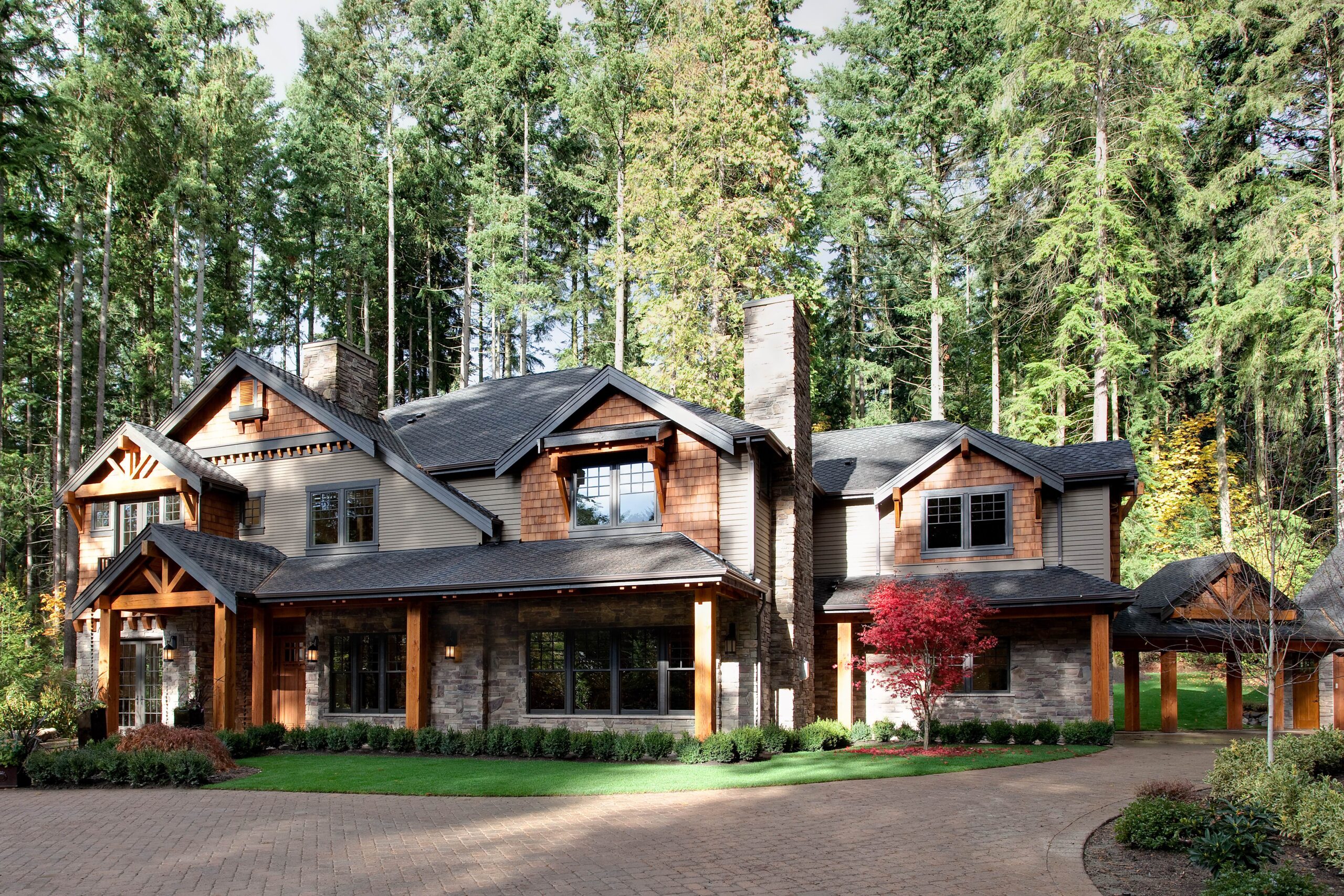 A large, modern two-story house with wood and stone exterior, surrounded by tall trees and lush greenery, featuring large windows, a front lawn, and a paved driveway.