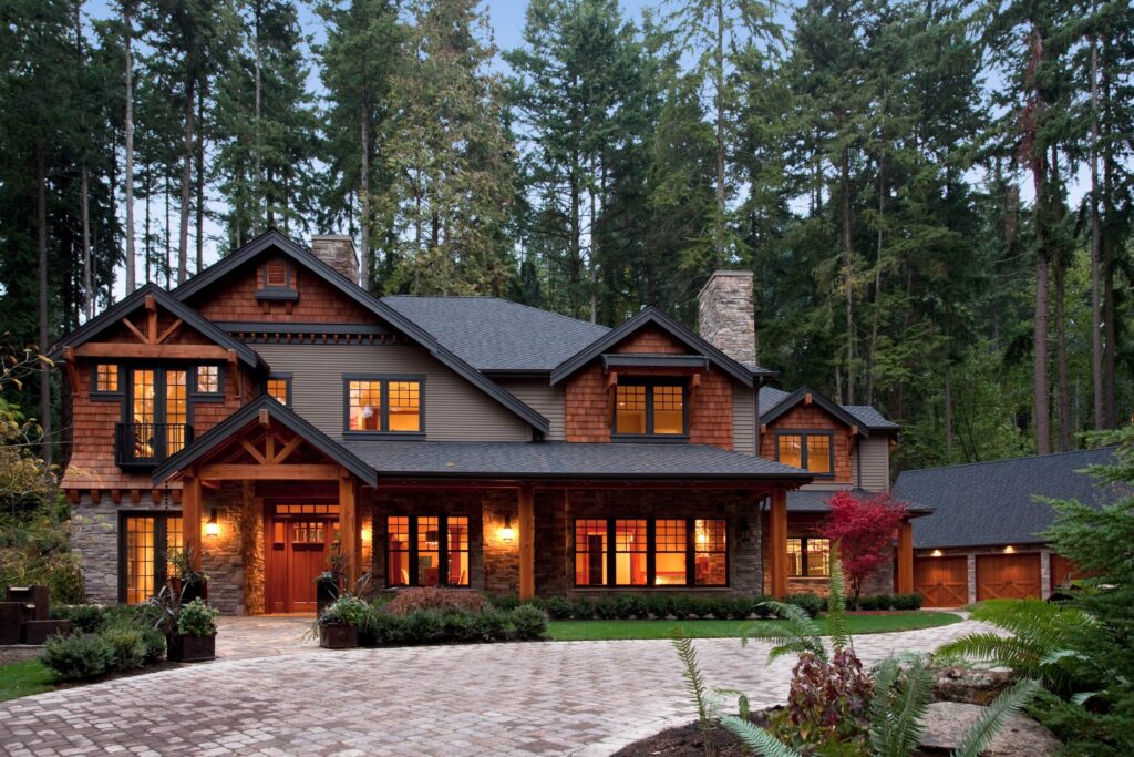 A large, two-story house with warm, glowing windows sits among tall trees. The home features wood and stone accents, a covered entryway, and a paved driveway leading to a detached garage. Lush greenery surrounds the property.
