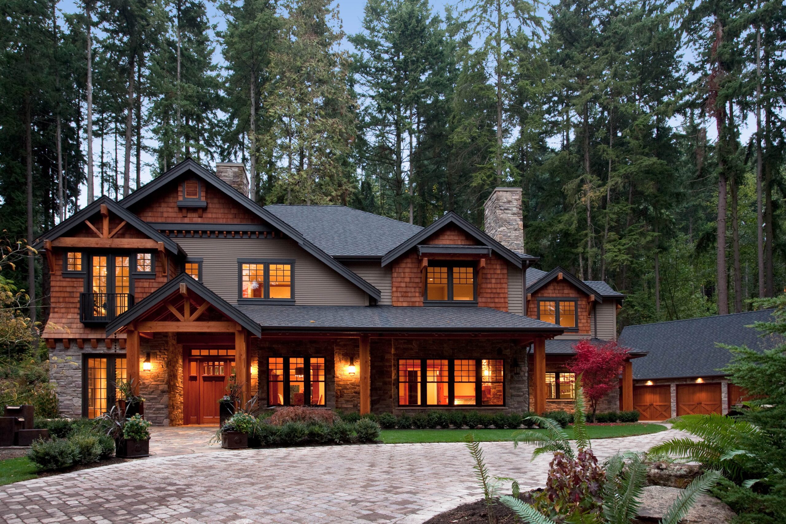 A large, two-story house with warm, glowing windows sits among tall trees. The home features wood and stone accents, a covered entryway, and a paved driveway leading to a detached garage. Lush greenery surrounds the property.