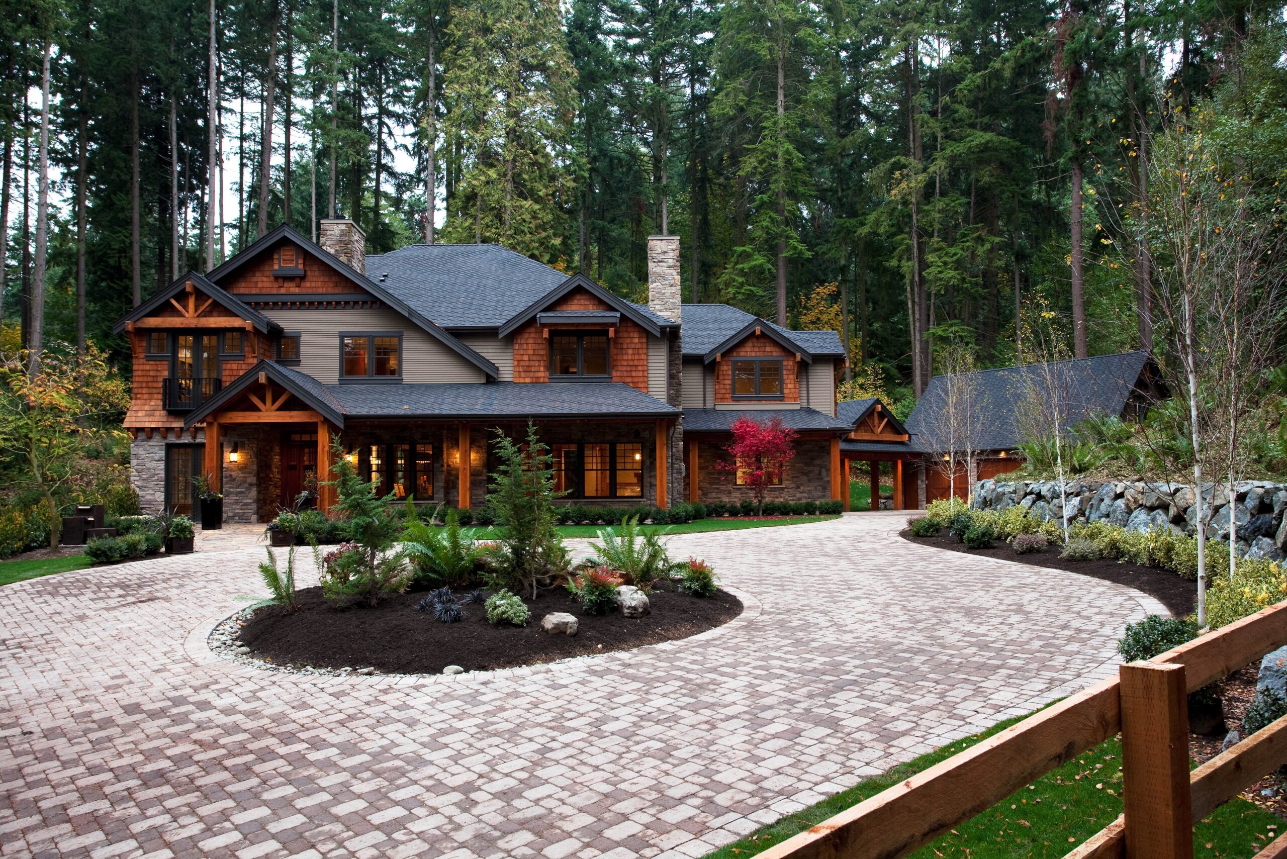 Large two-story house with wood and stone siding, surrounded by tall trees. A curved brick driveway leads to the entrance, landscaped with small bushes and trees. A detached garage is visible to the right.