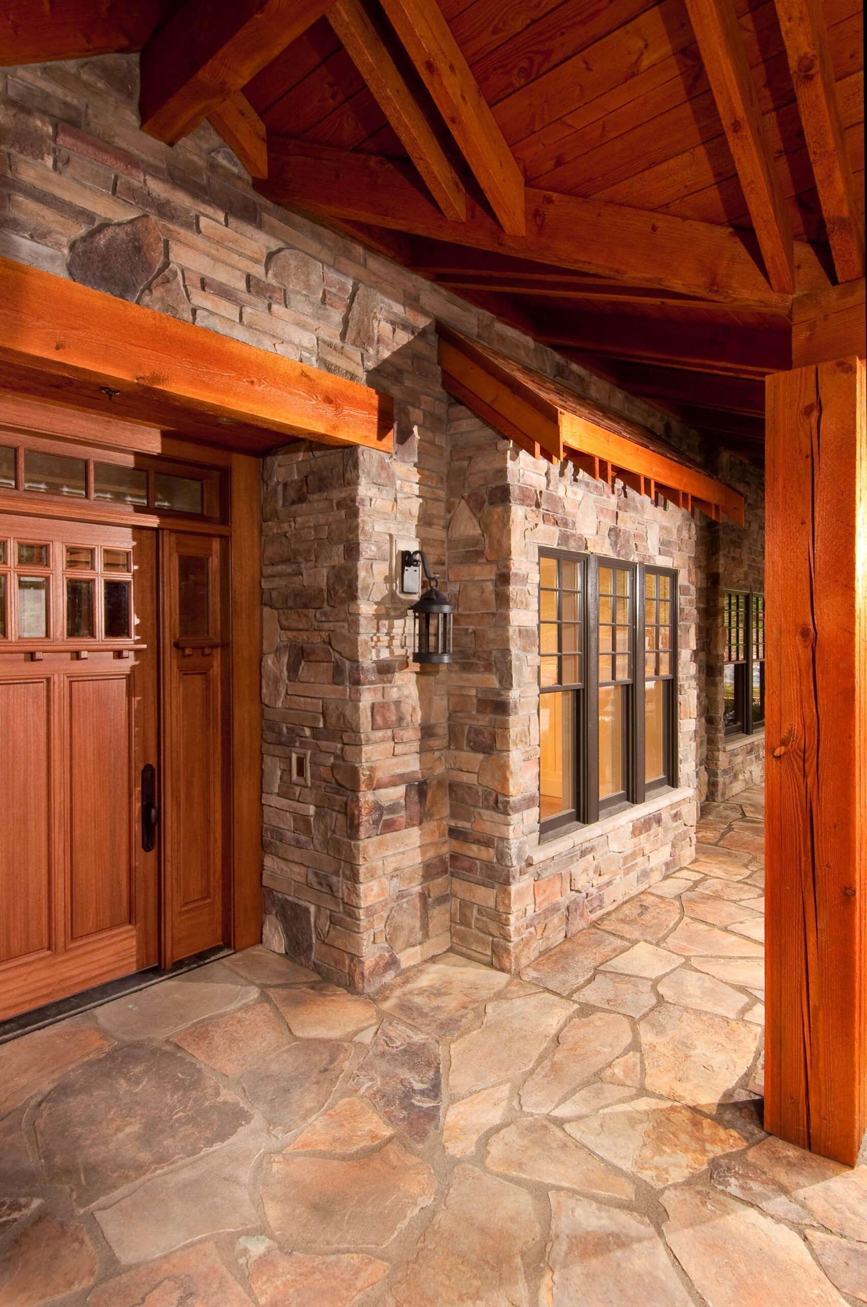 Covered porch with a wooden ceiling and beams, stone walls, multi-pane windows, and a wooden front door with glass panels and sidelights. The floor is made of irregular flagstone tiles.