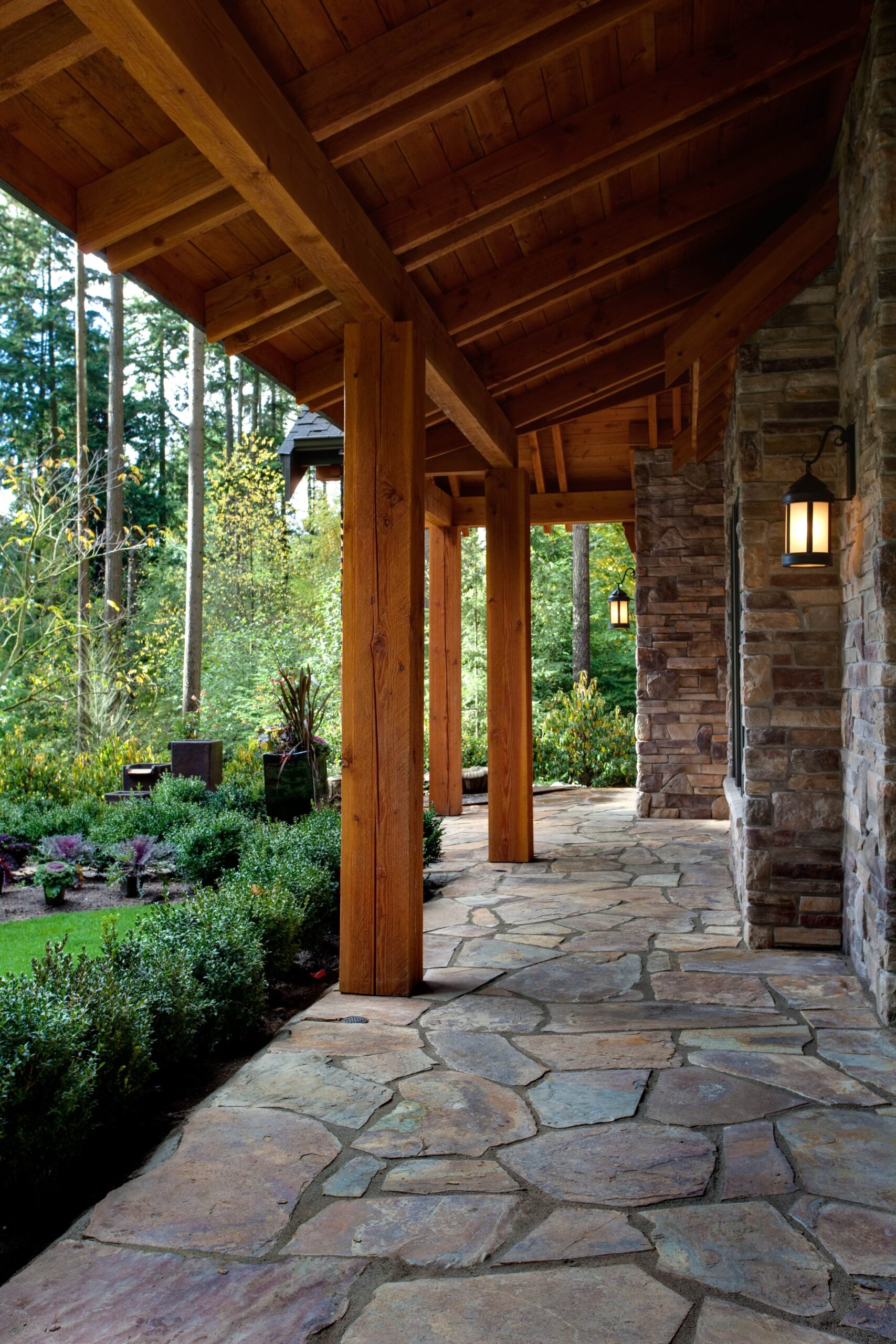 A stone pathway runs under a wooden porch roof supported by timber beams, adjacent to a stone wall with lantern-style lights. Lush green plants and tall trees surround the area.