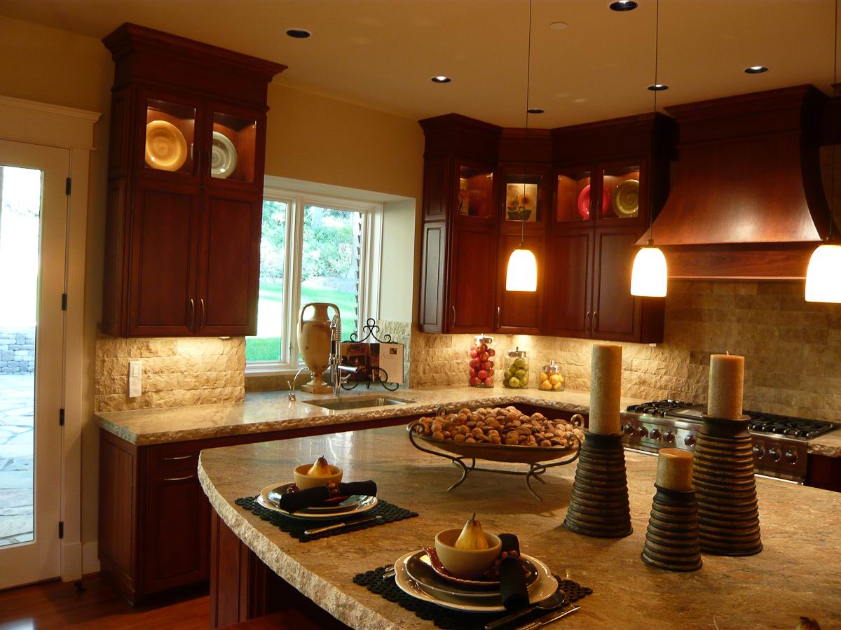 A warmly lit kitchen with dark wooden cabinets, granite countertops, pendant lights, decorative candles, plates, and bowls on the counter, and a window overlooking a green outdoor area.