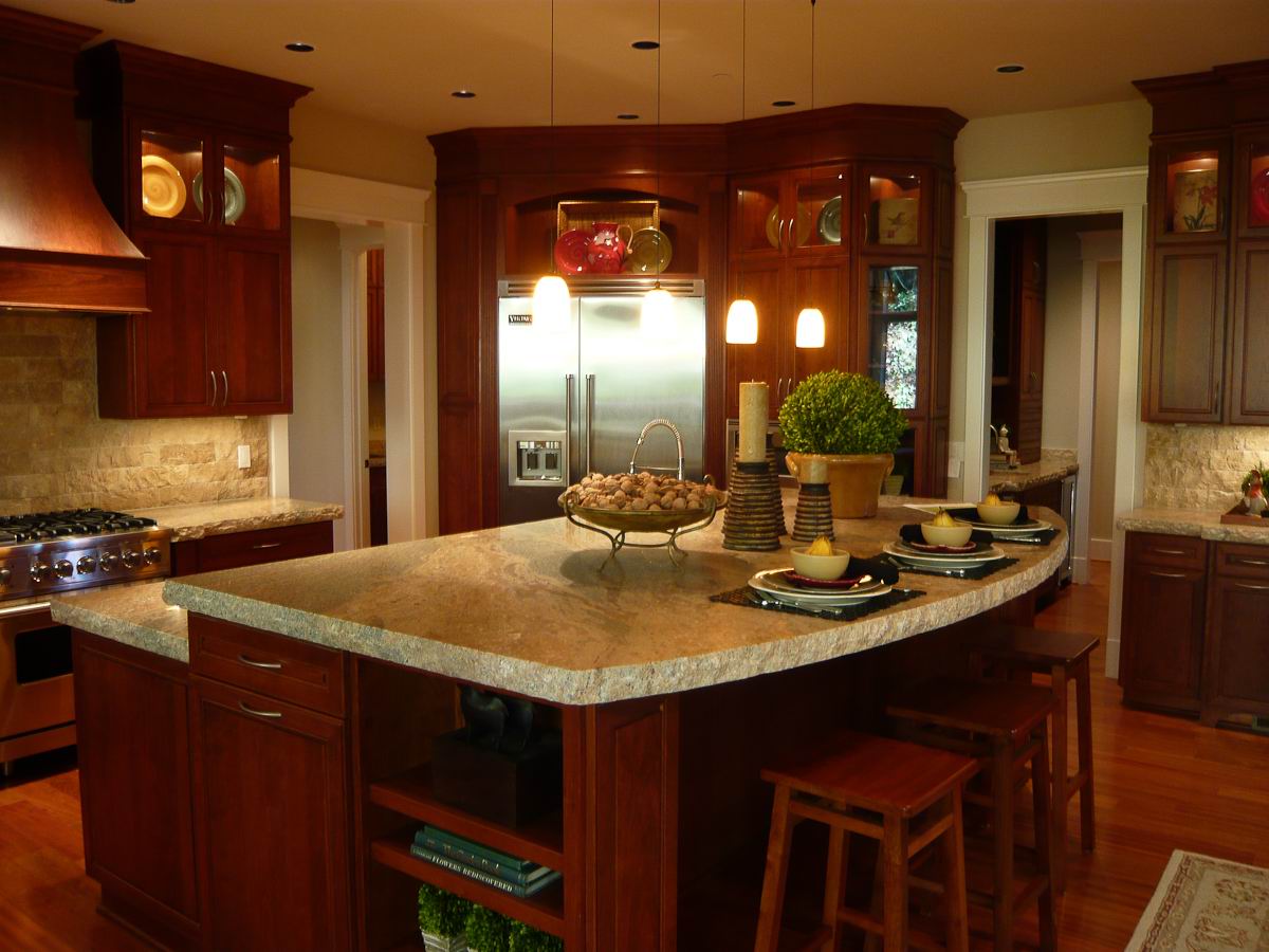A spacious kitchen with dark wood cabinets, stainless steel appliances, and a large island with a marble countertop. The island has stools, place settings, potted plants, and decorative bowls. Warm lighting creates a cozy atmosphere.
