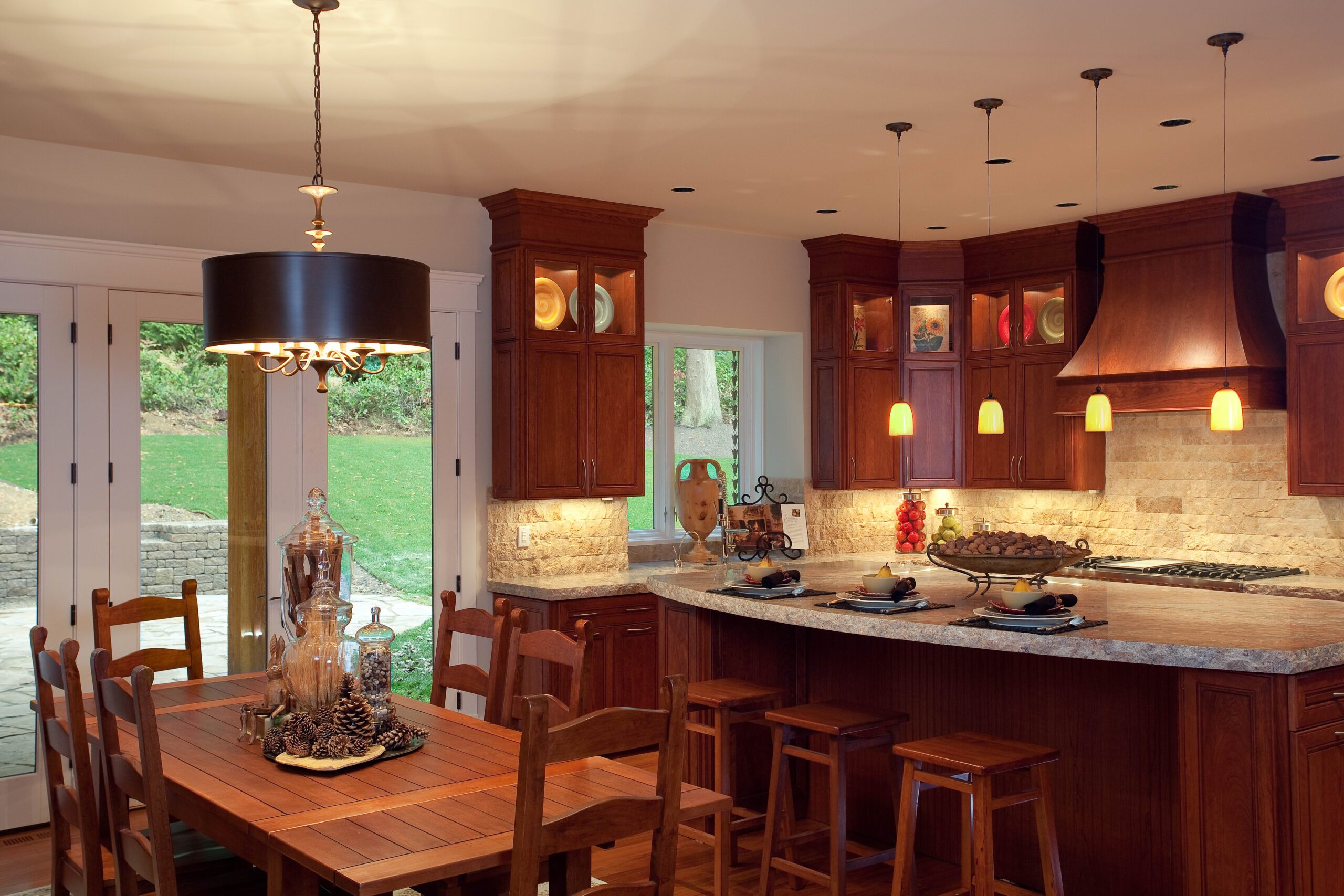 A warm, inviting kitchen with wooden cabinets, a large island with barstools, pendant lights, and a dining table set for four. Sliding glass doors open to a green backyard, letting in natural light.