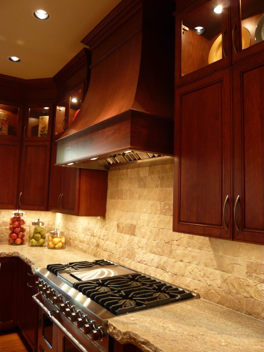 A kitchen with dark wood cabinets, a stainless steel gas stove, a stone backsplash, and glass jars filled with fruit on the granite countertop under warm lighting.
