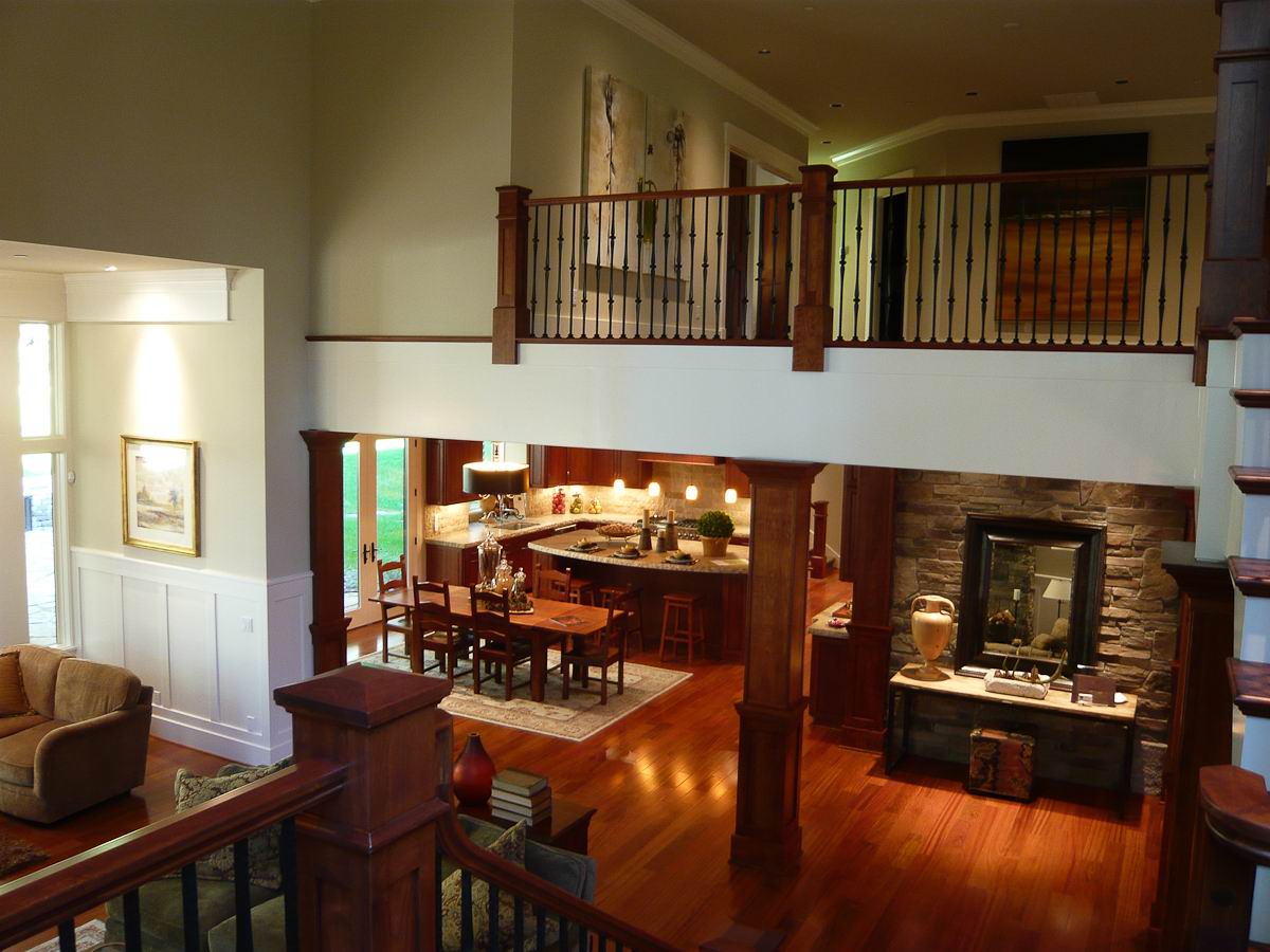 A spacious, elegant open-concept living area with high ceilings, wooden floors, a stone fireplace, and a central kitchen and dining space. Upstairs, a balcony overlooks the main floor. Warm lighting creates a cozy atmosphere.