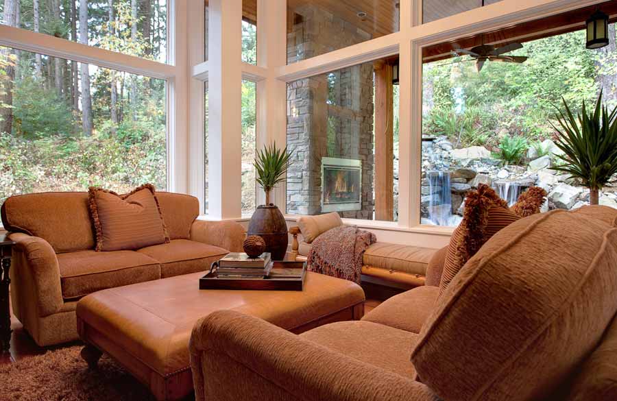 Cozy living room with tan sofas and ottoman, large windows with views of trees and a waterfall, potted plants, a fireplace outside, and a decorative tray on the ottoman. Natural light fills the space.