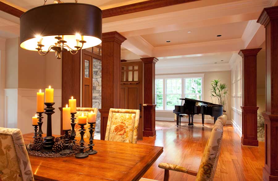 A dining room with a wooden table, patterned chairs, a black chandelier, and lit candles. In the background, a grand piano sits by large windows, letting in natural light. The space features wooden floors and columns.