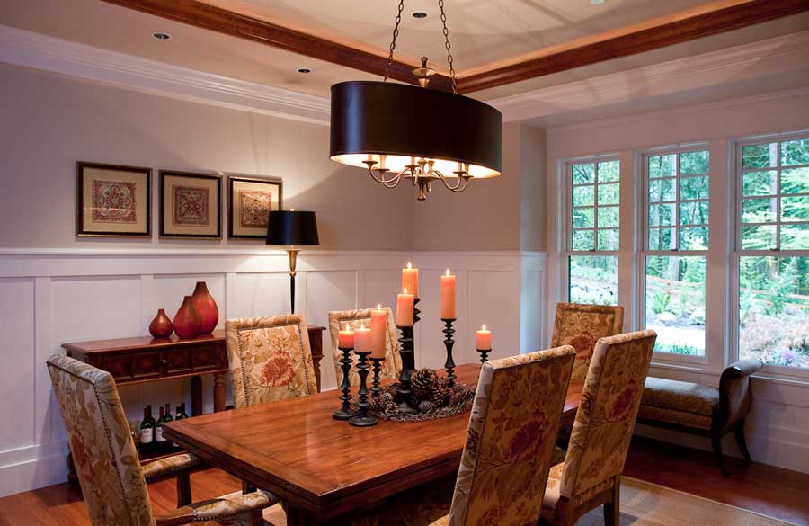 A cozy dining room with a wooden table, six upholstered chairs, a black chandelier, and candle centerpieces. A window seat, framed art, and decorative vases enhance the warm, inviting atmosphere.