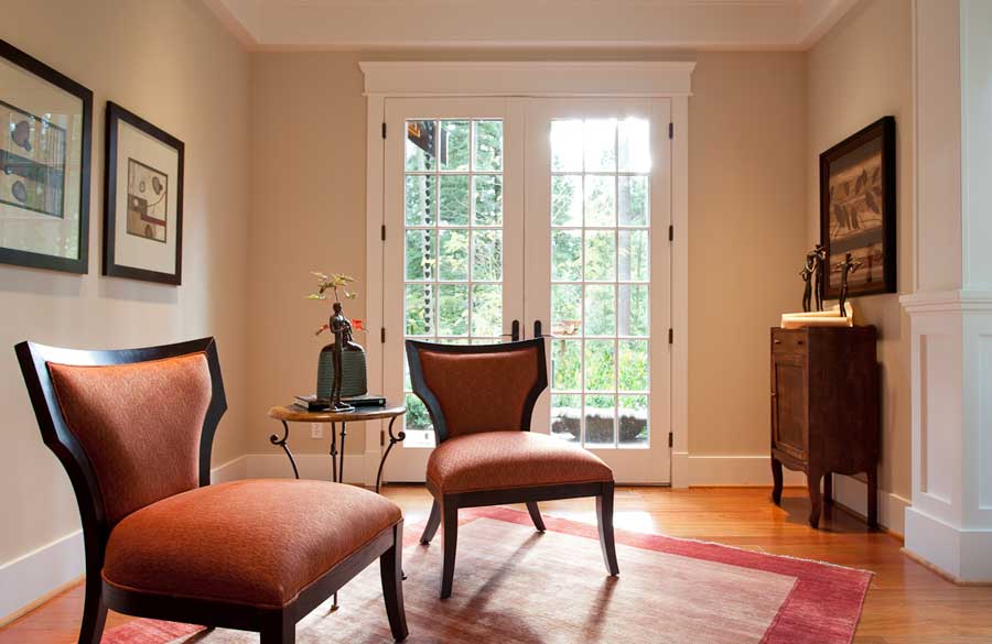A cozy sitting area with two orange upholstered chairs, a small side table, and framed art on the walls. Double glass doors let in natural light, highlighting a red rug and wooden floor. A cabinet stands against the far wall.