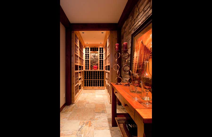 A warmly lit wine cellar with wooden shelves filled with bottles, stone walls, and a table holding candles, glasses, and a framed picture along the right side of the hallway.