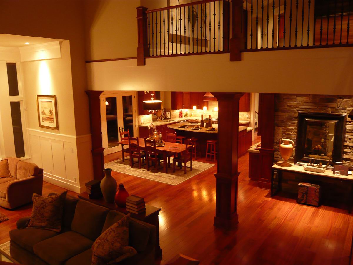 Warmly lit, open-concept living room and kitchen with wooden floors, a dining table, cushioned sofas, decorative vases, stone fireplace, and an upstairs railing overlooking the space.