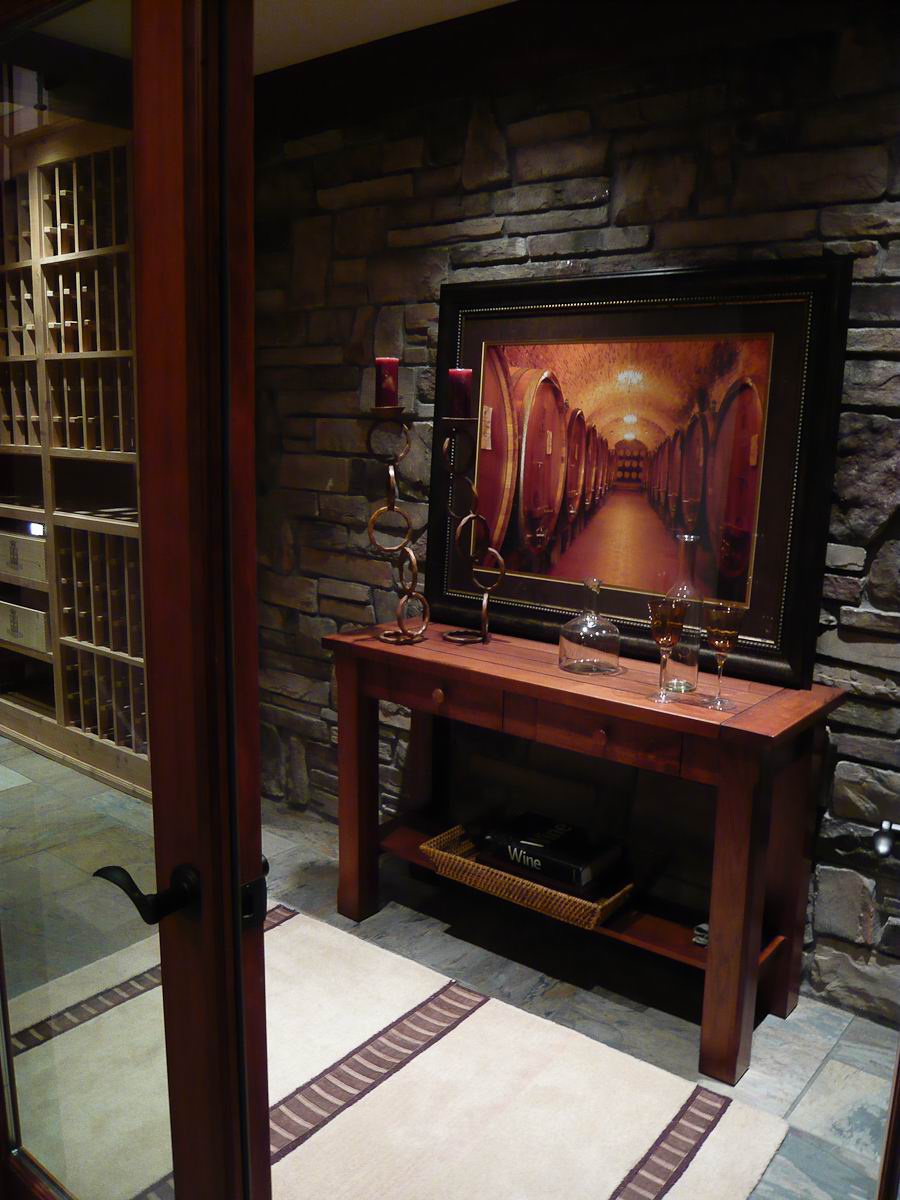 A wine cellar entry with stone walls, a wooden table holding glassware and candles, and a large framed picture of wine barrels. Wine storage racks are visible through a glass door on the left.