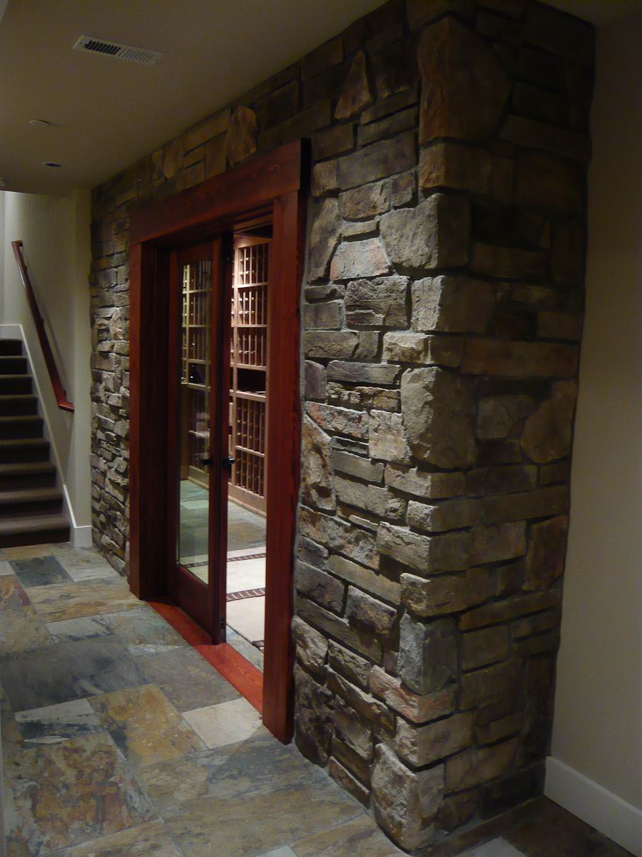 A stone wall with a wooden-framed glass door leads to a wine cellar. The floor is made of large stone tiles, and a carpeted staircase is visible on the left side of the image.