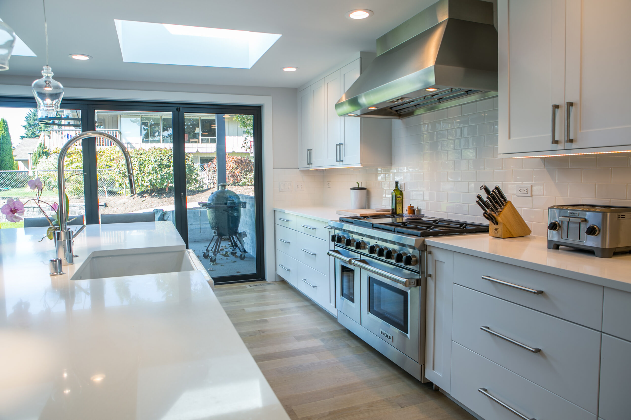 Modern kitchen with white cabinets, stainless steel appliances, a large stove, and a toaster. There’s a sink island, knife block, and sliding glass doors leading to a patio with a barbecue grill outside. Natural light fills the room.