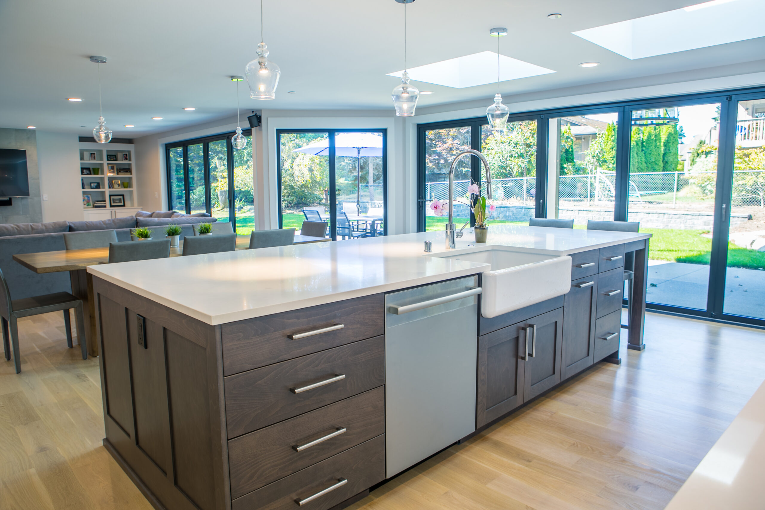 Modern kitchen with a large island featuring a farmhouse sink, stainless steel dishwasher, pendant lights, and floor-to-ceiling windows offering a view of a patio and greenery outside.
