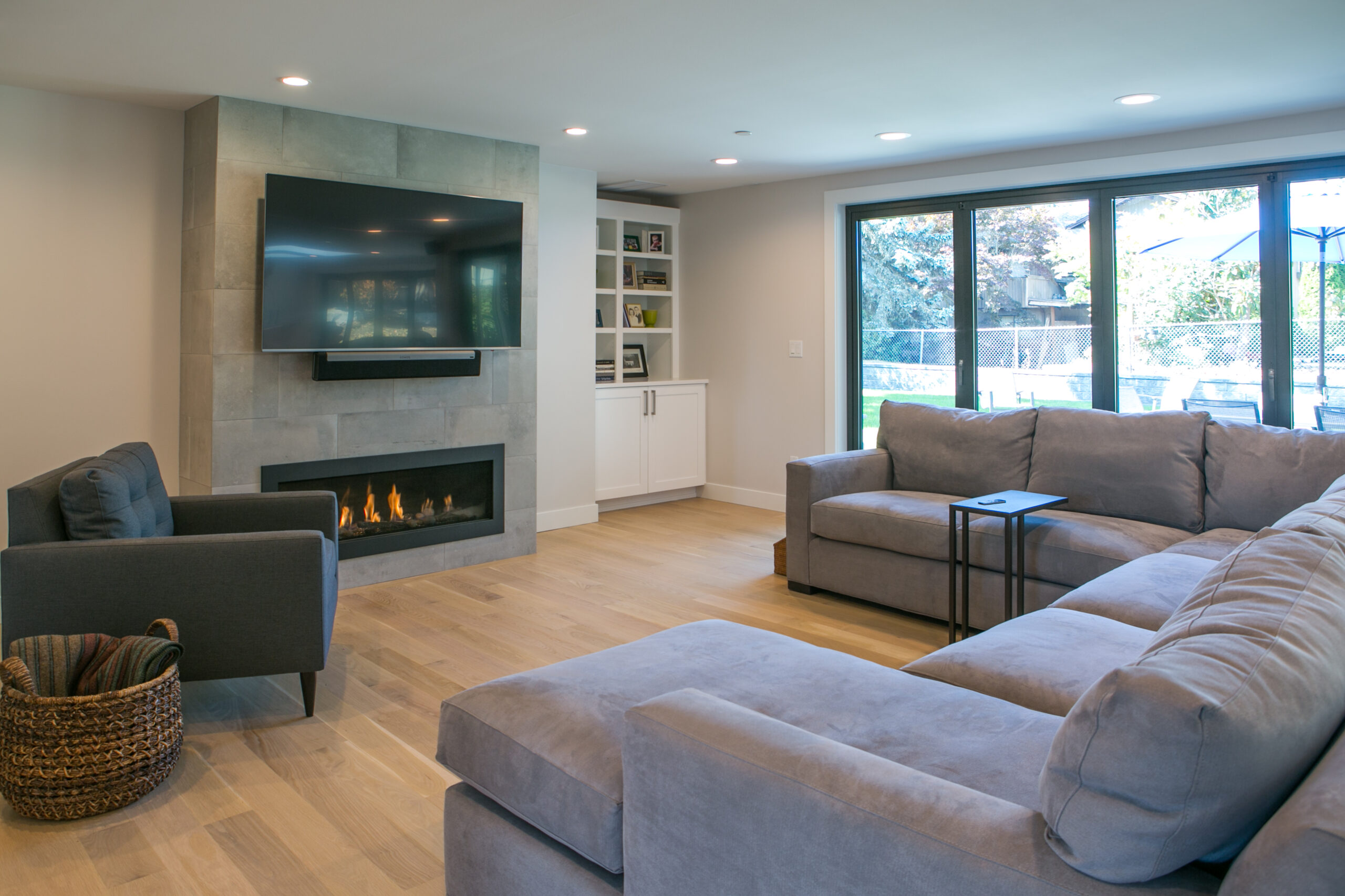 Modern living room with large gray sectional sofa, armchair, wall-mounted TV above a fireplace, built-in shelves, light wood flooring, and glass doors opening to a backyard. A woven basket is beside the armchair.
