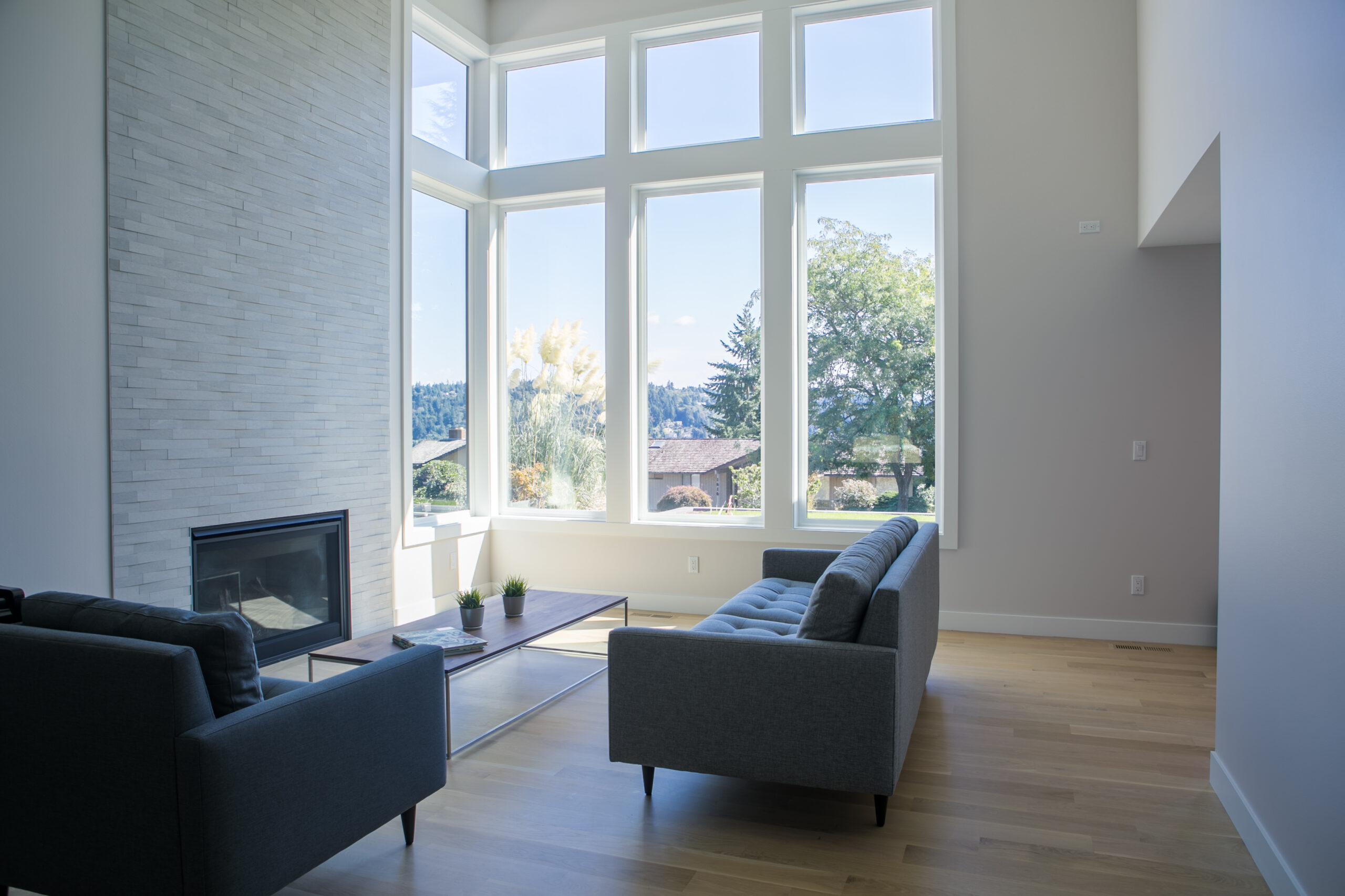 Modern living room with large floor-to-ceiling windows, two gray sofas, a glass coffee table with small plants, light wood flooring, and a fireplace with a white textured wall. Trees and sky are visible outside.