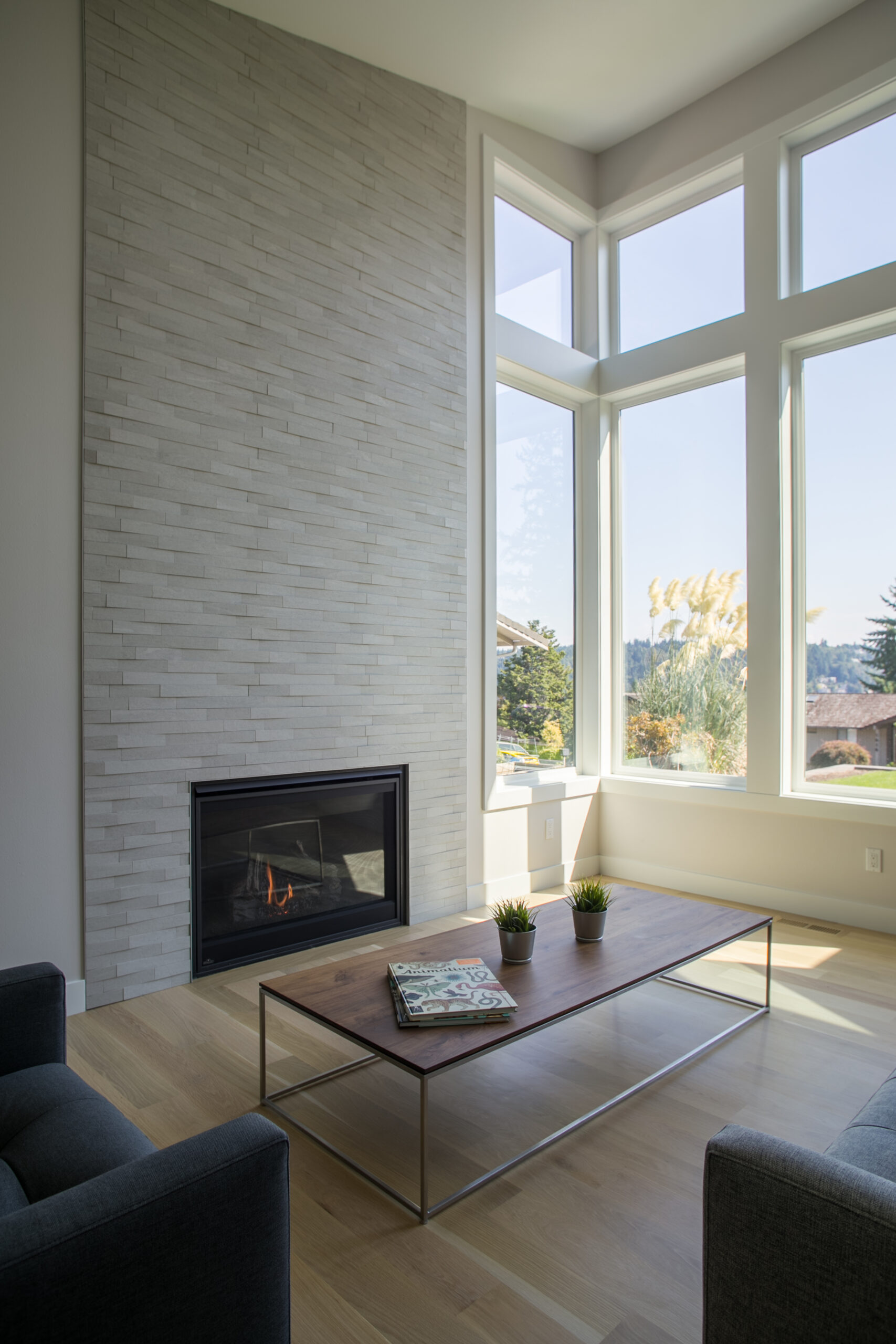 Modern living room with a tall stone fireplace, large floor-to-ceiling windows letting in natural light, a wooden coffee table with small potted plants and magazines, and a light wood floor.