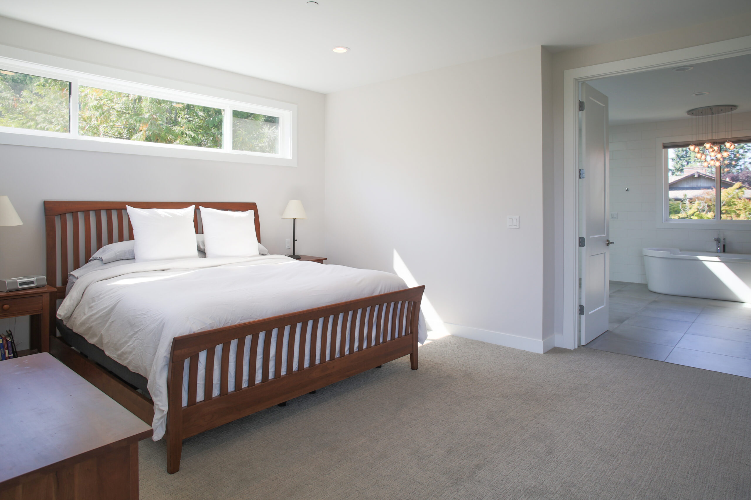 A bright, modern bedroom with a wooden bed frame, white bedding, two bedside tables with lamps, beige carpet, a large window, and an open door leading to a bathroom with a bathtub.