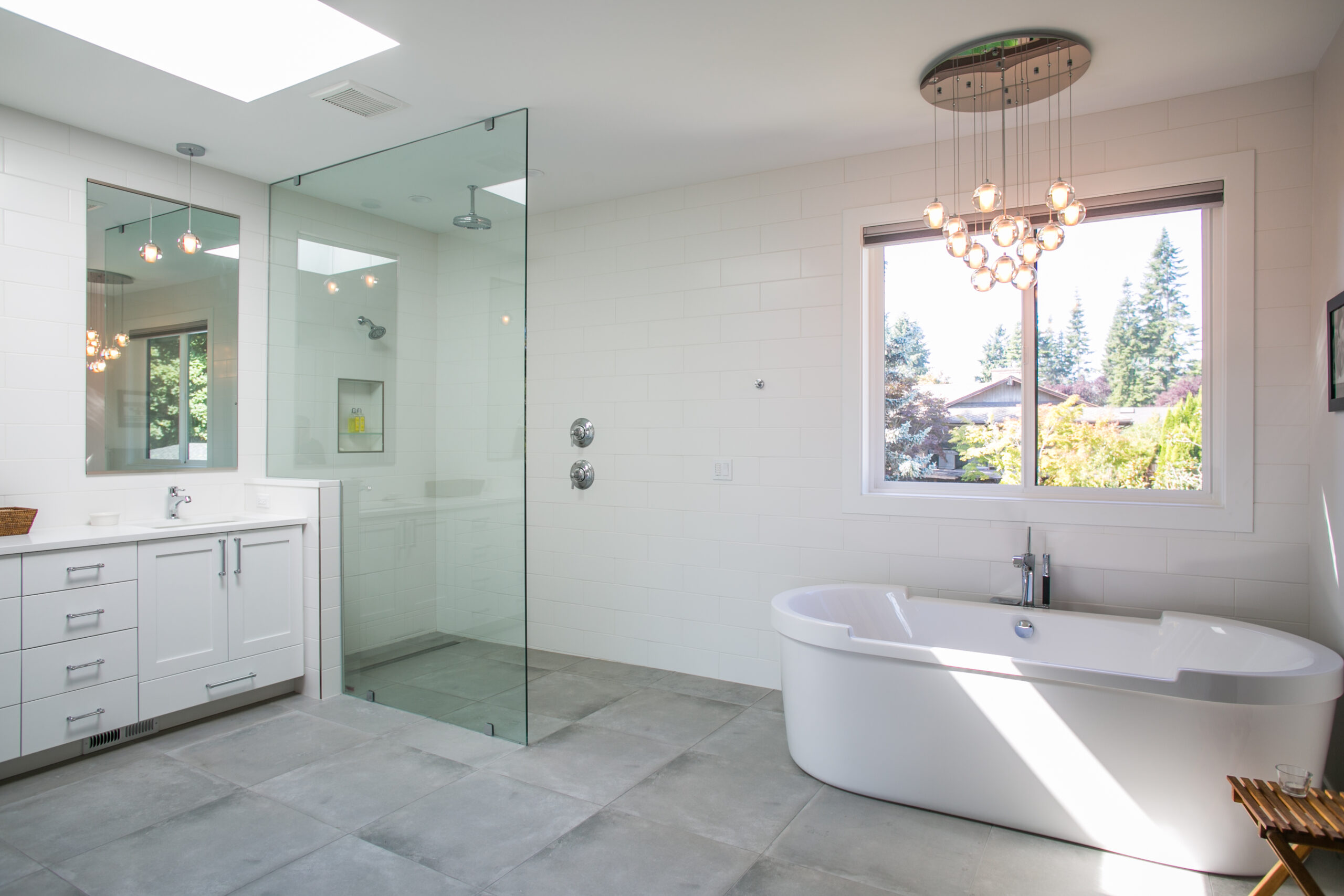 A modern, bright bathroom with a freestanding bathtub, large glass-enclosed shower, white vanity, tile flooring, and a window overlooking trees. A chandelier hangs from the ceiling.