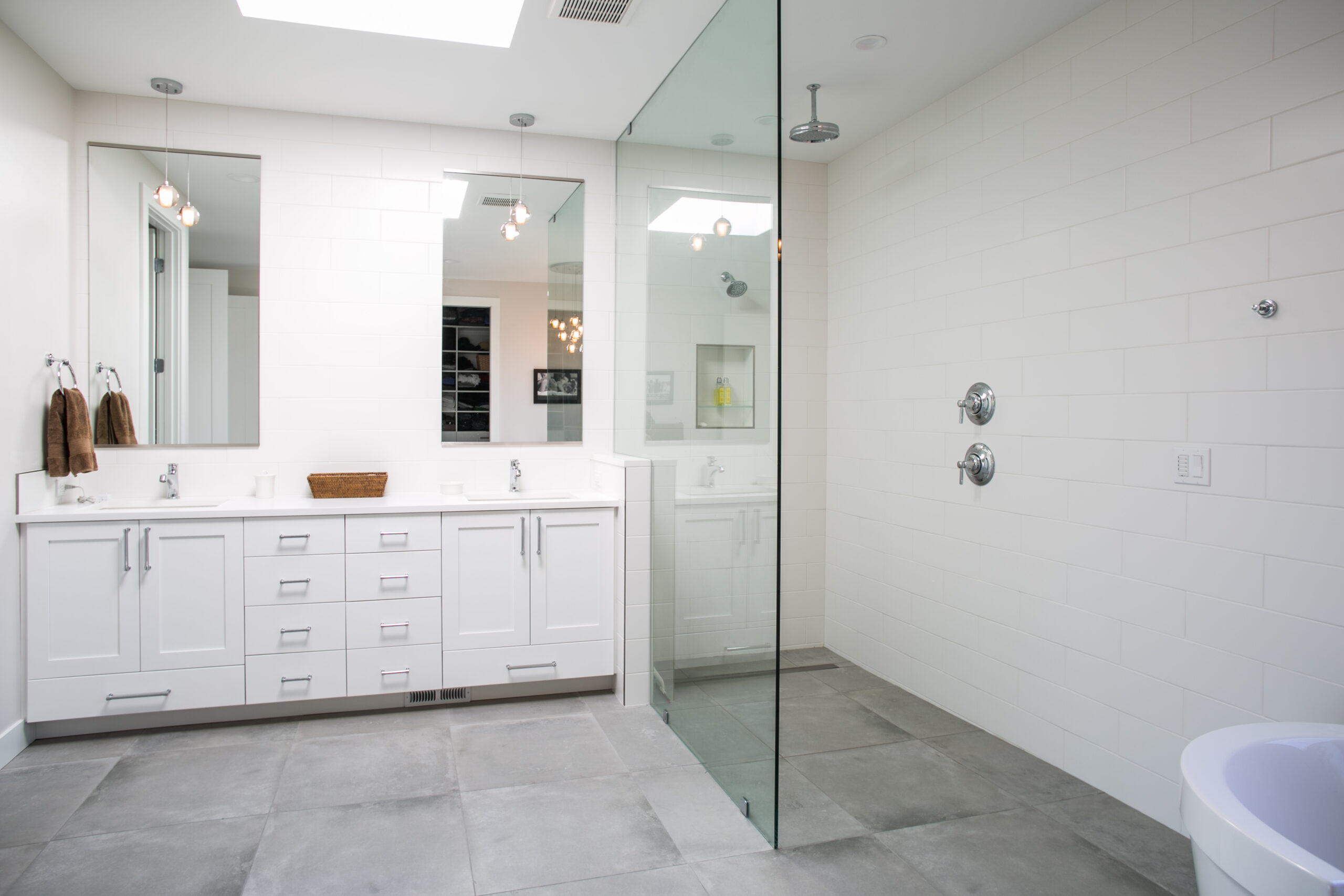 Modern bathroom with a double-sink white vanity, large mirrors, towels, and a spacious walk-in shower enclosed by glass. The room features white walls, gray tile floors, and minimalist decor.