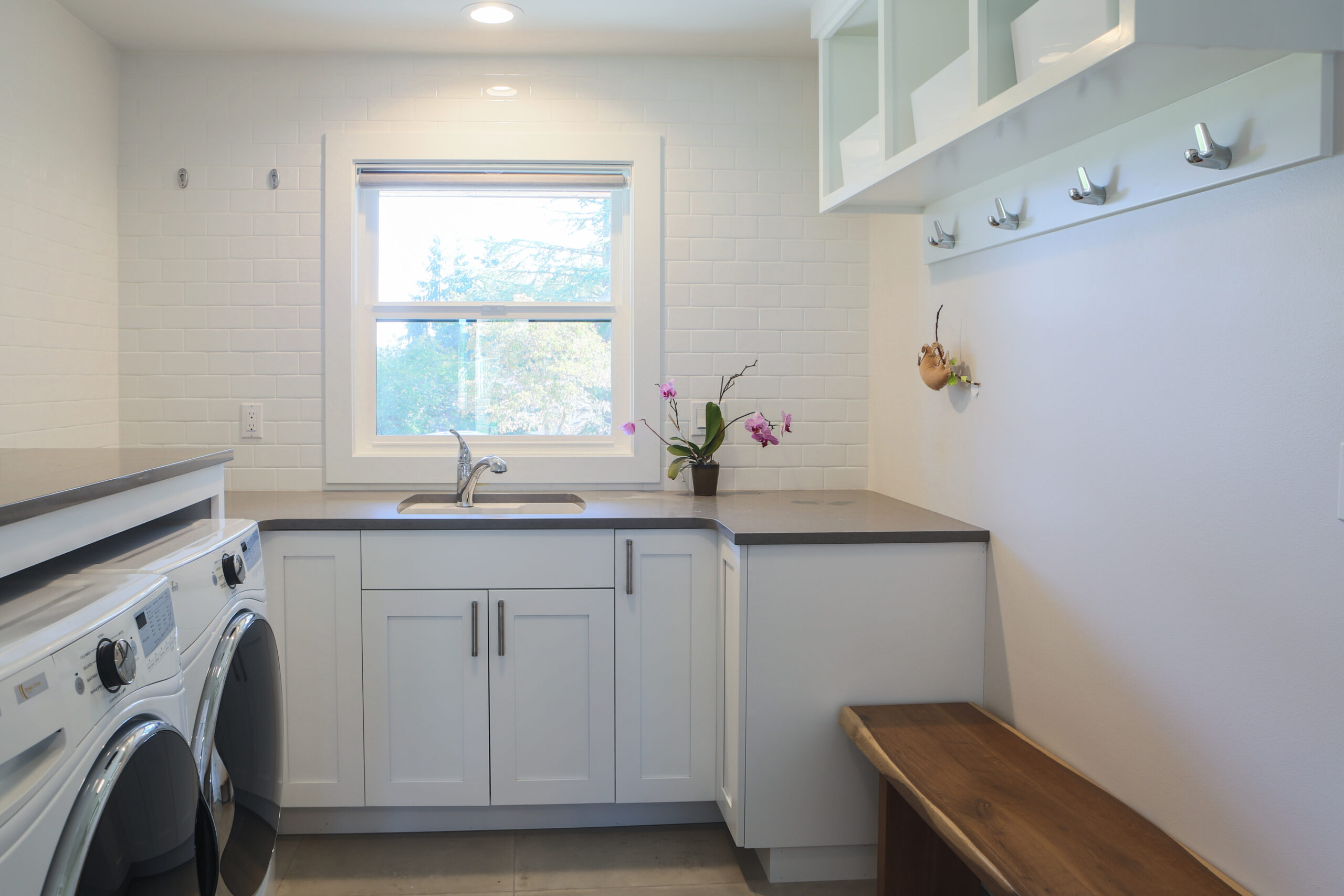 Bright laundry room with white cabinets, a front-loading washer and dryer, a window above a sink, wall hooks, a wooden bench, and a small potted orchid on the countertop.