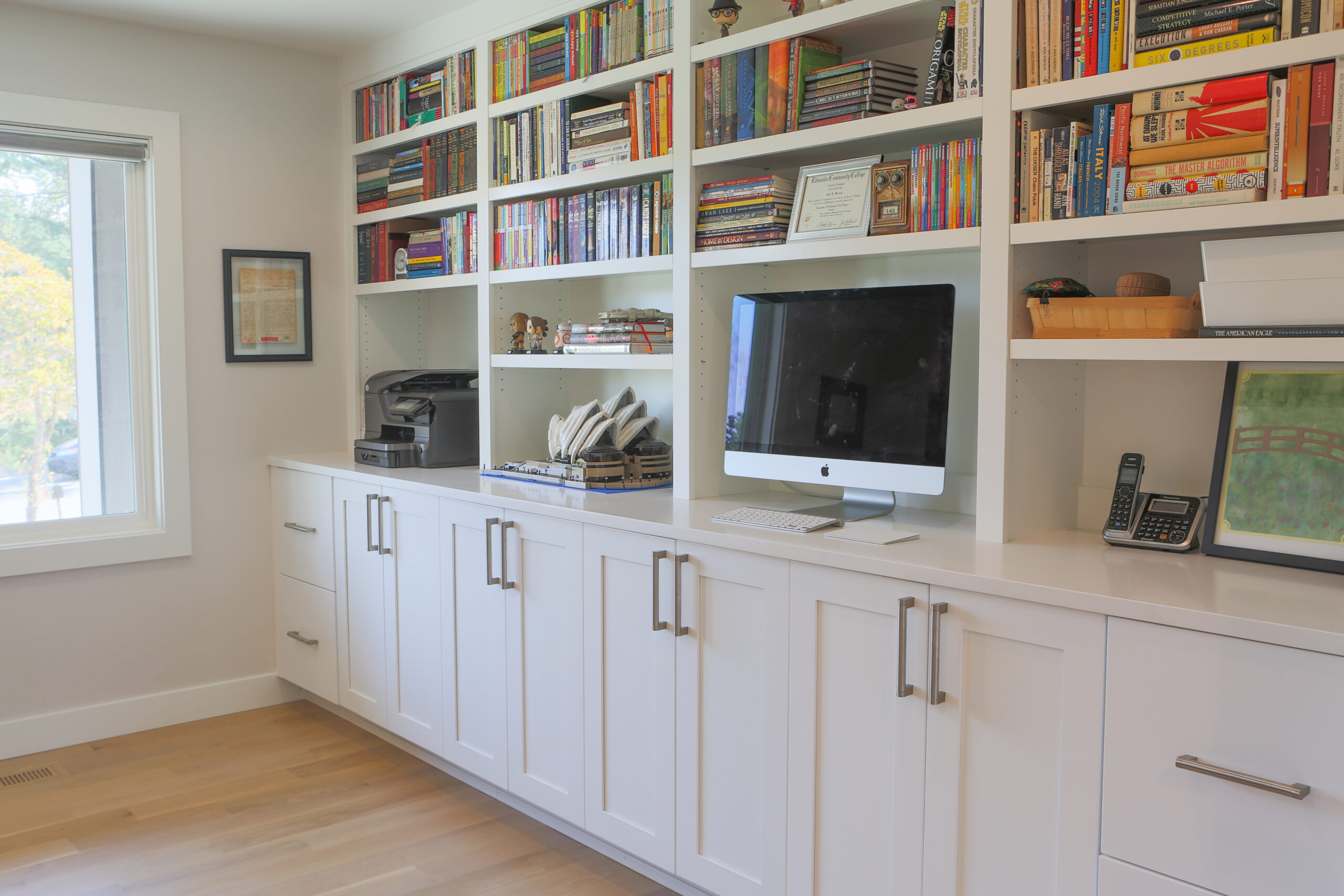 A modern home office features white cabinets and shelves filled with books and decor. A desktop computer, printer, and office supplies sit on the countertop beneath the shelves. Natural light enters through a nearby window.