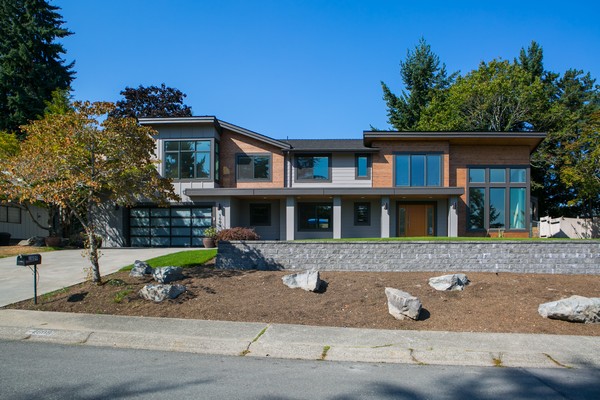Modern two-story house with large windows, a mix of wood and gray siding, and a flat roof. The front yard has rocks, sparse plants, and a driveway leading to a glass garage door. Trees surround the property.