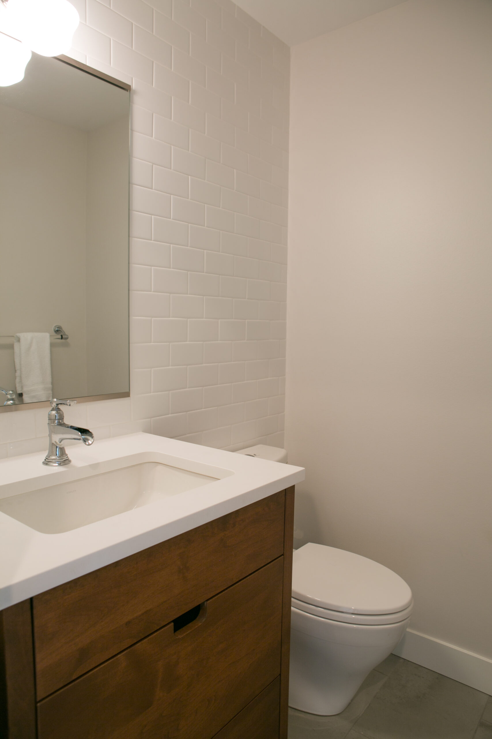 A modern bathroom with a wooden vanity, white countertop and sink, chrome faucet, wall mirror, white subway tile backsplash, and a white toilet next to the vanity.