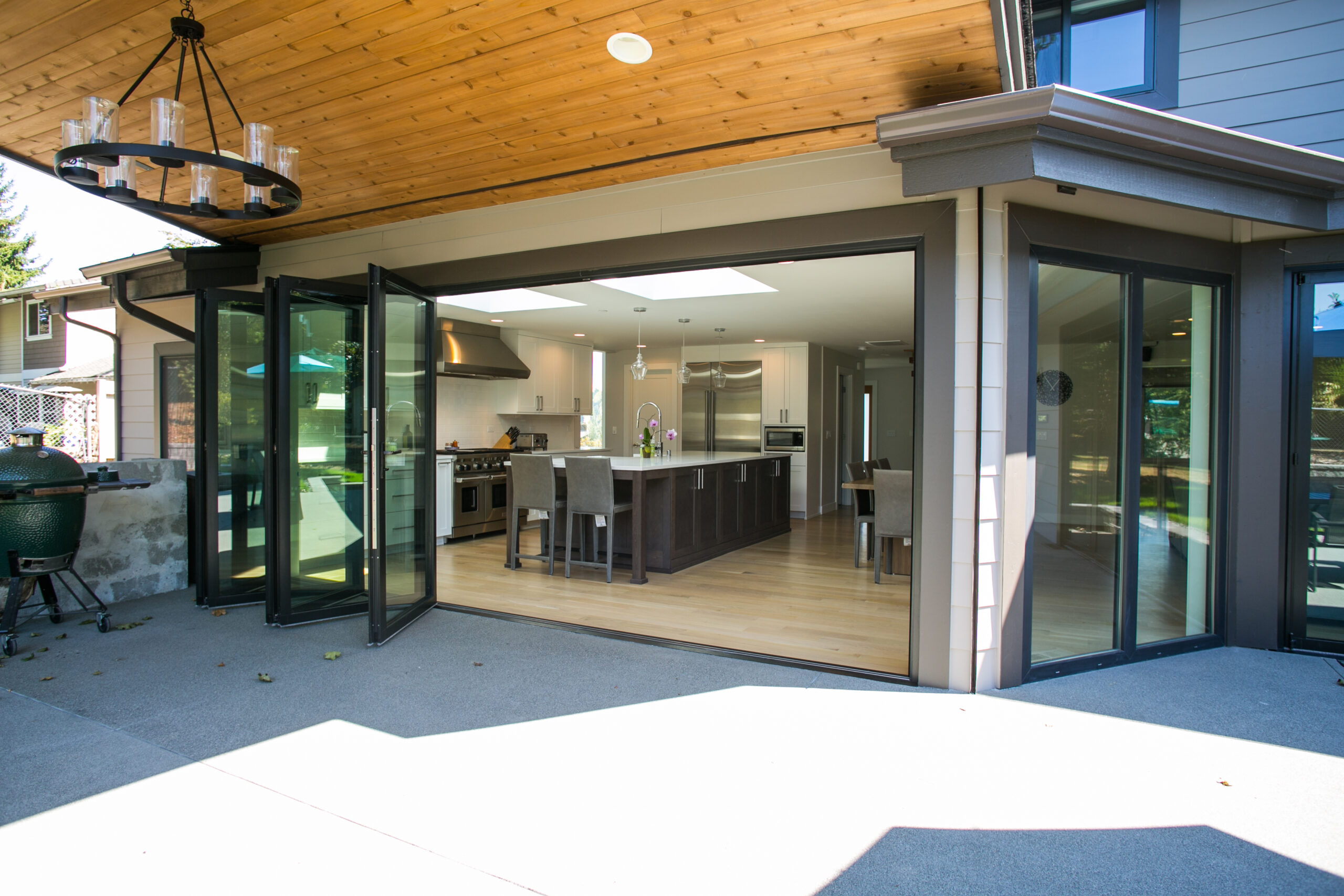 Modern home with large folding glass doors open to a spacious kitchen and dining area, featuring wood floors, a central island with barstools, stainless steel appliances, and a wood-paneled ceiling on the patio.