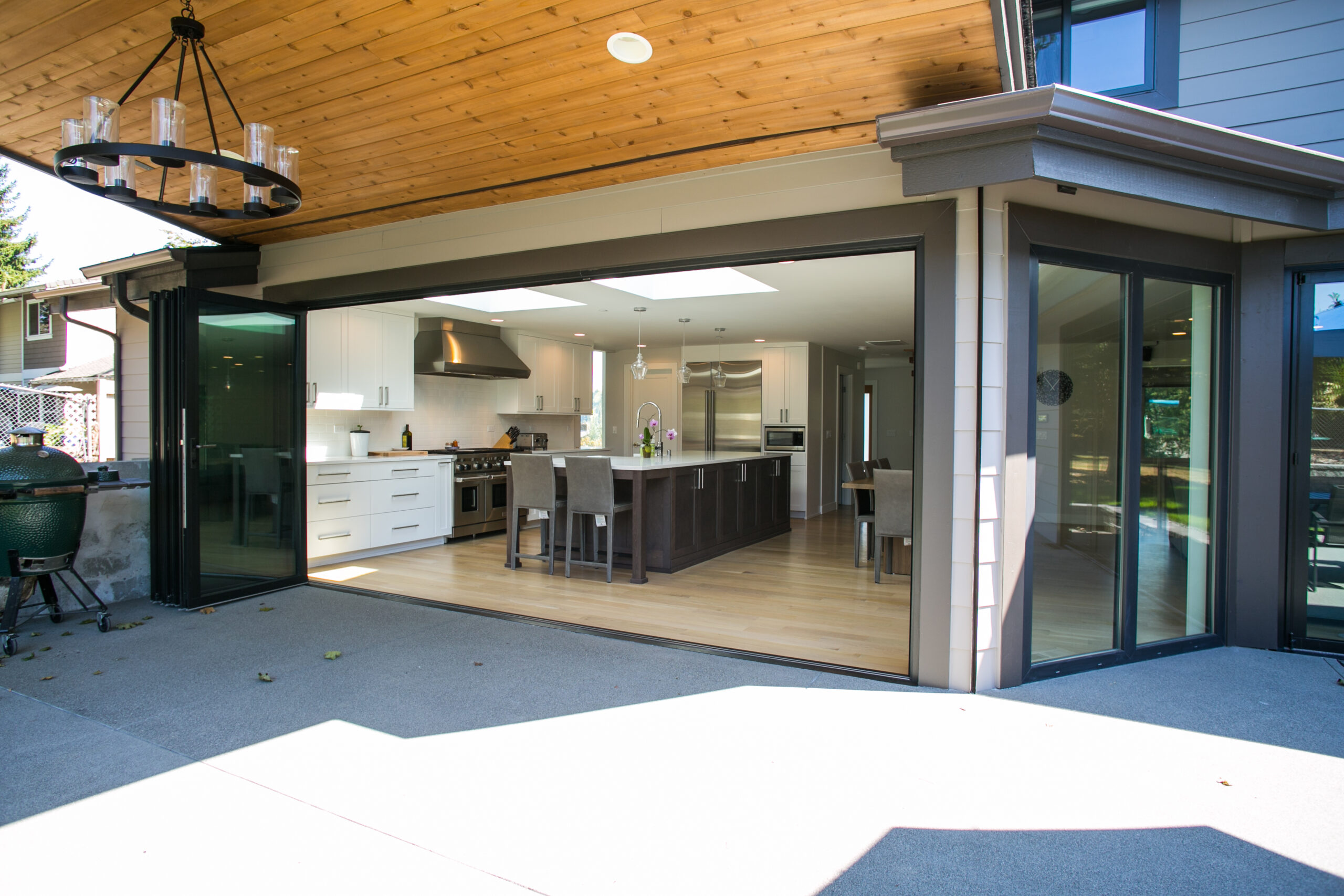 A modern kitchen and dining area with white cabinets, stainless steel appliances, and a large island, viewed through wide open glass sliding doors leading to a covered outdoor patio.