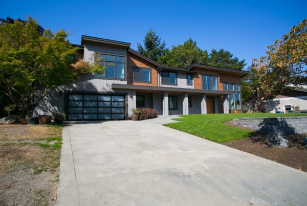 Modern two-story house with large windows, a double garage, and a spacious driveway, surrounded by green grass, trees, and landscaping under a clear blue sky.