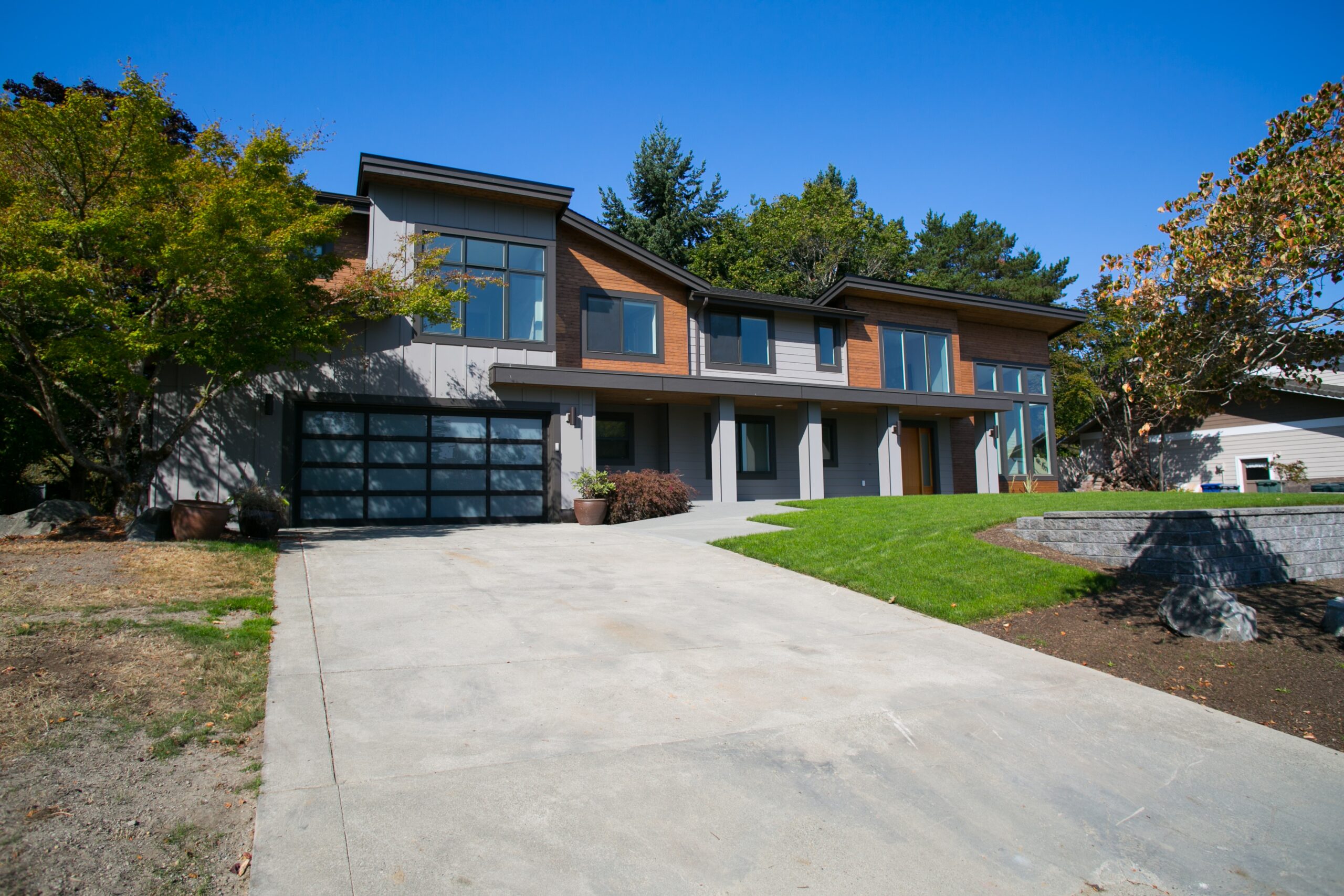 Modern two-story house with large windows, a double garage, and a spacious driveway, surrounded by green grass, trees, and landscaping under a clear blue sky.
