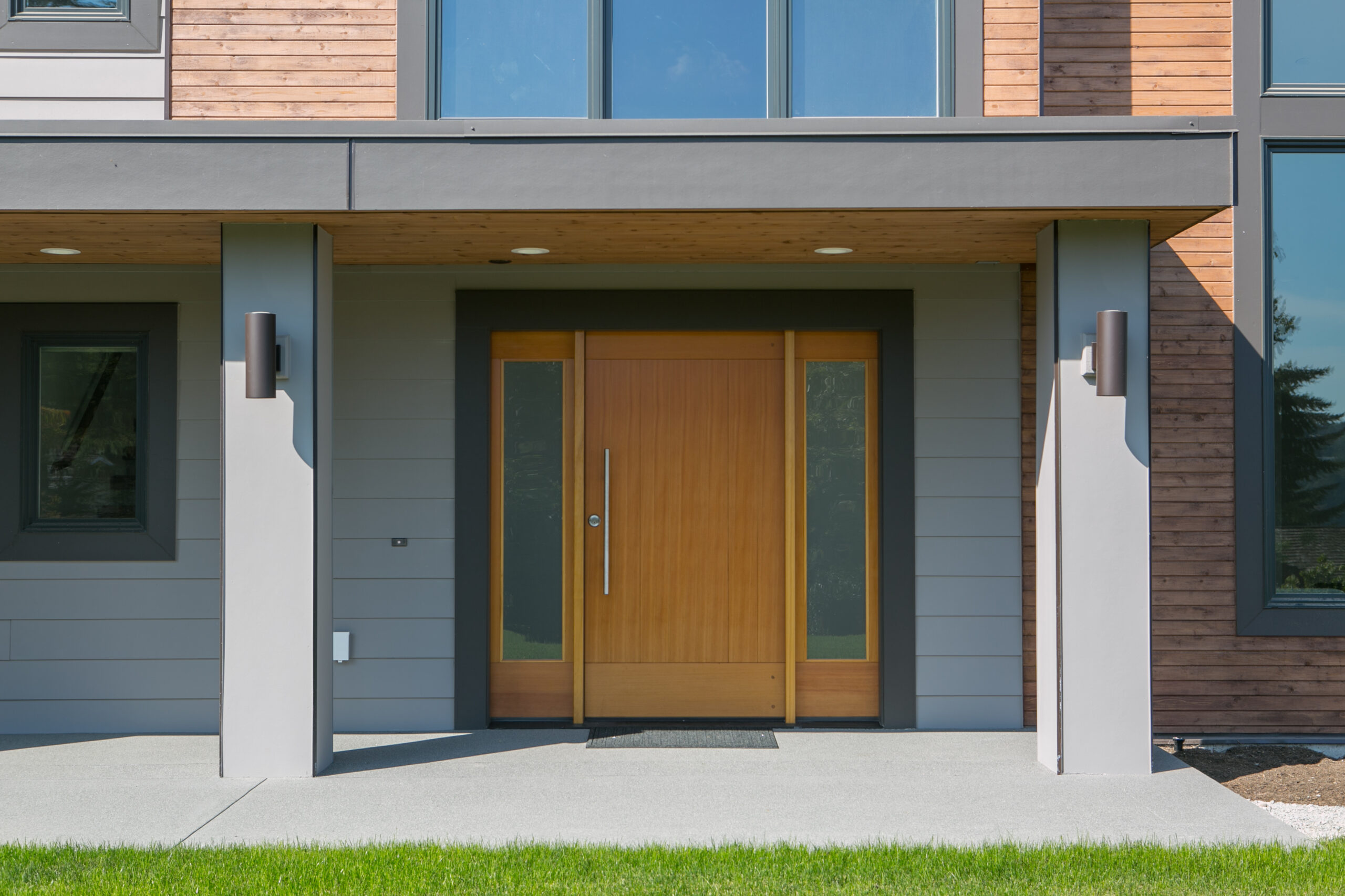 Modern house entrance with a large wooden front door flanked by two narrow frosted glass panels, gray siding, wood accents, and two wall-mounted outdoor lights. Green lawn in foreground.