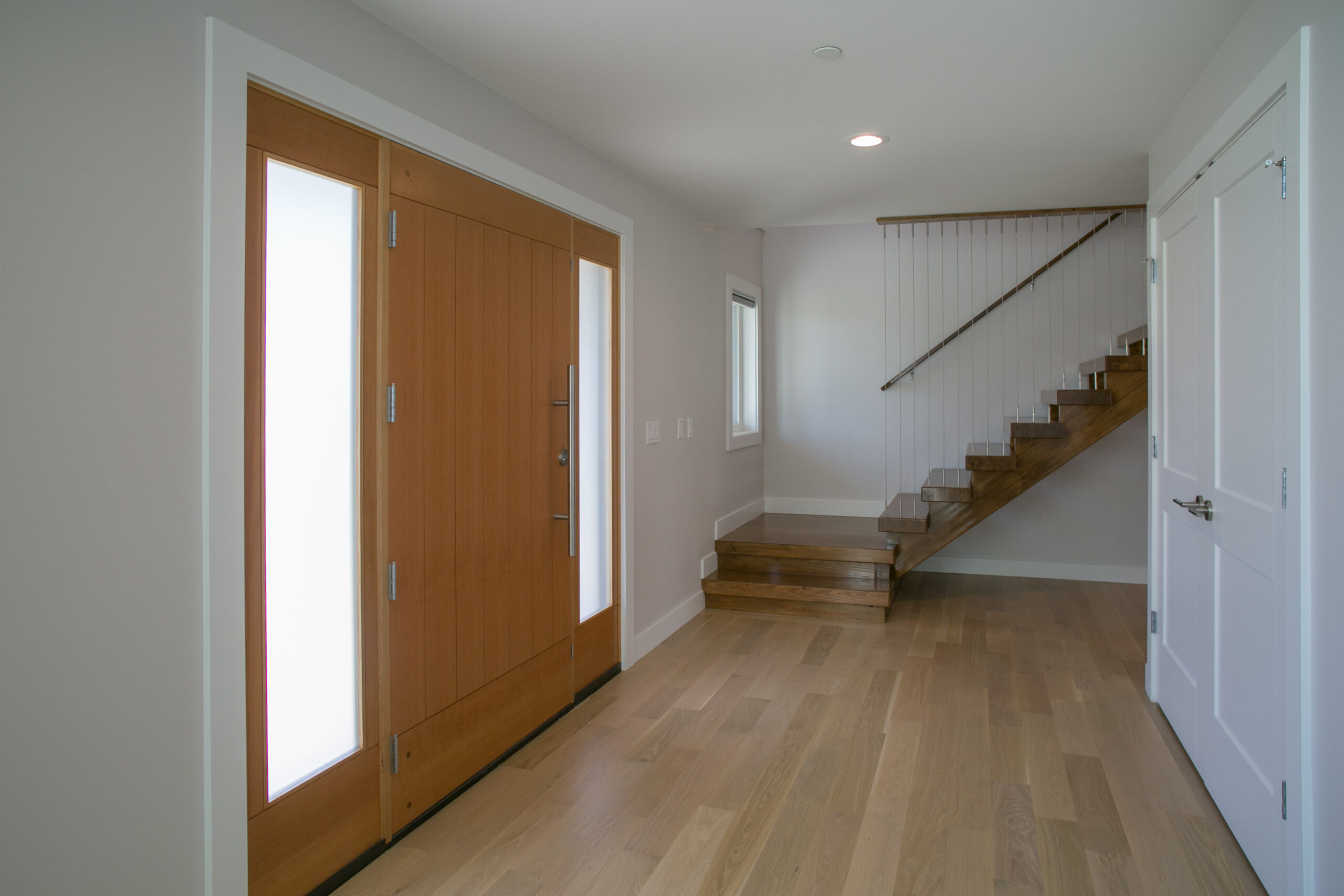 A modern entryway with light wood floors, a wooden front door with frosted glass panels, white walls, and a wooden staircase with metal railings leading up to the second floor.