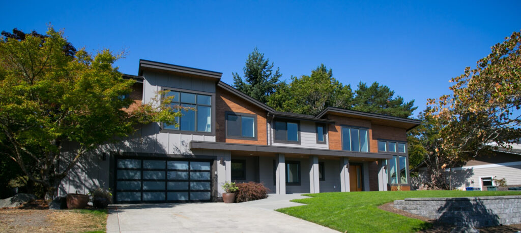 Modern two-story house with large glass windows, wood and gray siding, a double garage, and landscaped front yard with green grass and trees under a clear blue sky.