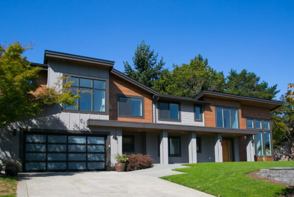 Modern two-story house with large glass windows, wood and gray siding, a double garage, and landscaped front yard with green grass and trees under a clear blue sky.