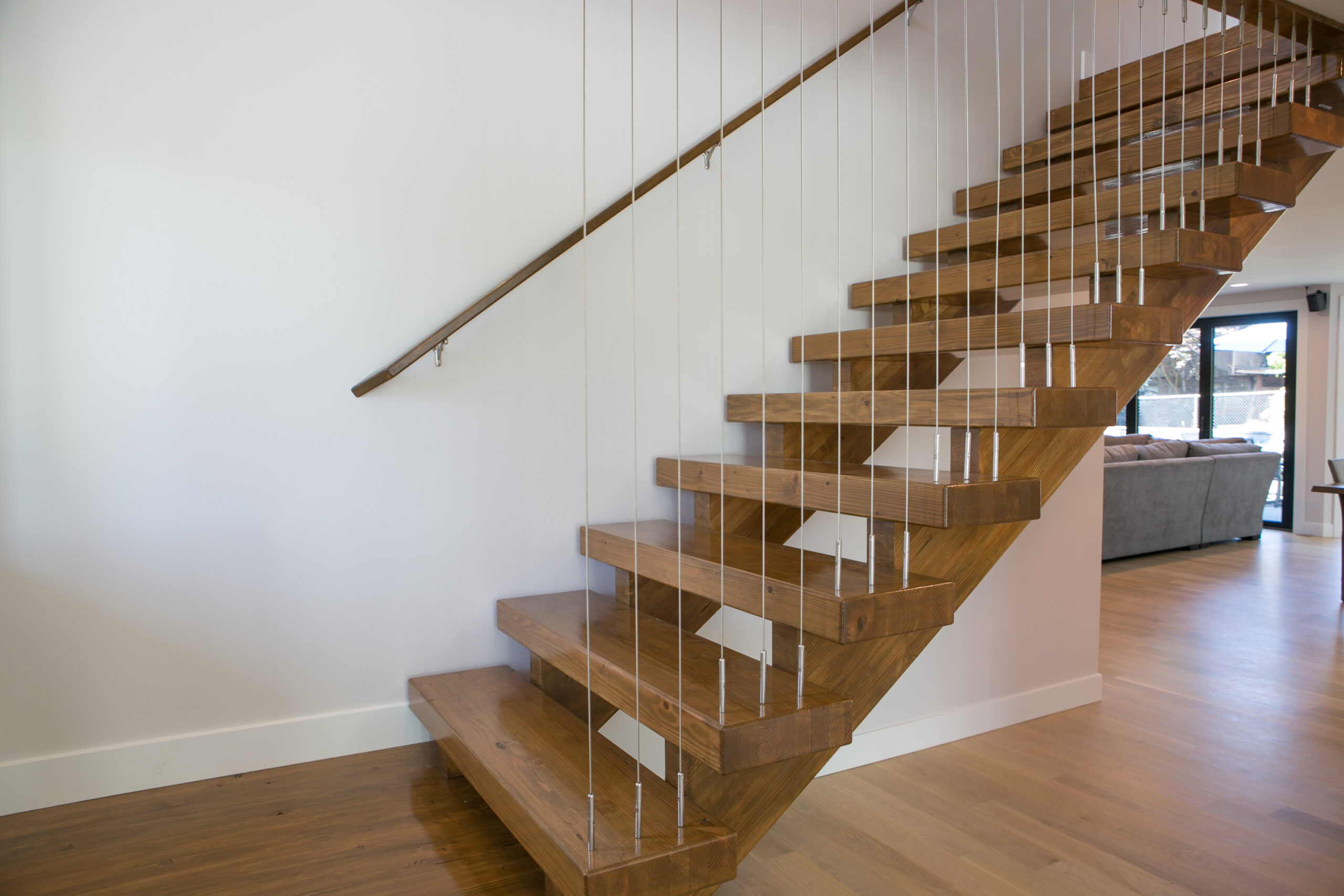 Modern wooden staircase with open risers and metal cables serving as a railing, ascending alongside a white wall in a bright, contemporary interior with hardwood floors.