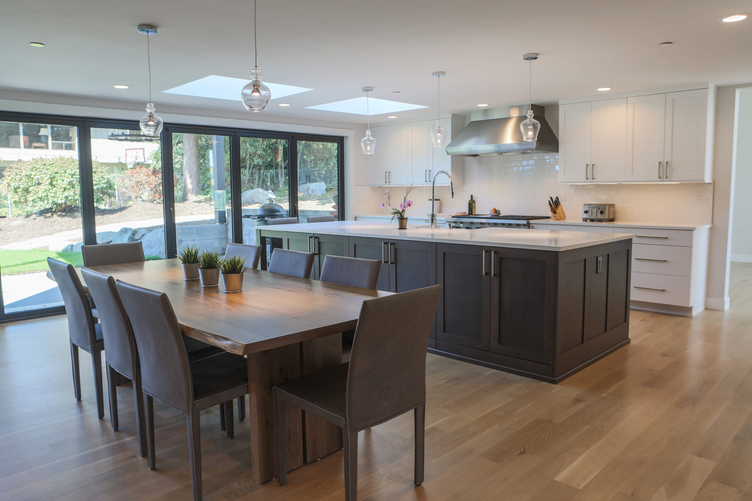 Modern kitchen and dining area with a wooden table and six chairs, large island with dark cabinets, white countertops, stainless steel appliances, pendant lights, and sliding glass doors opening to a garden.
