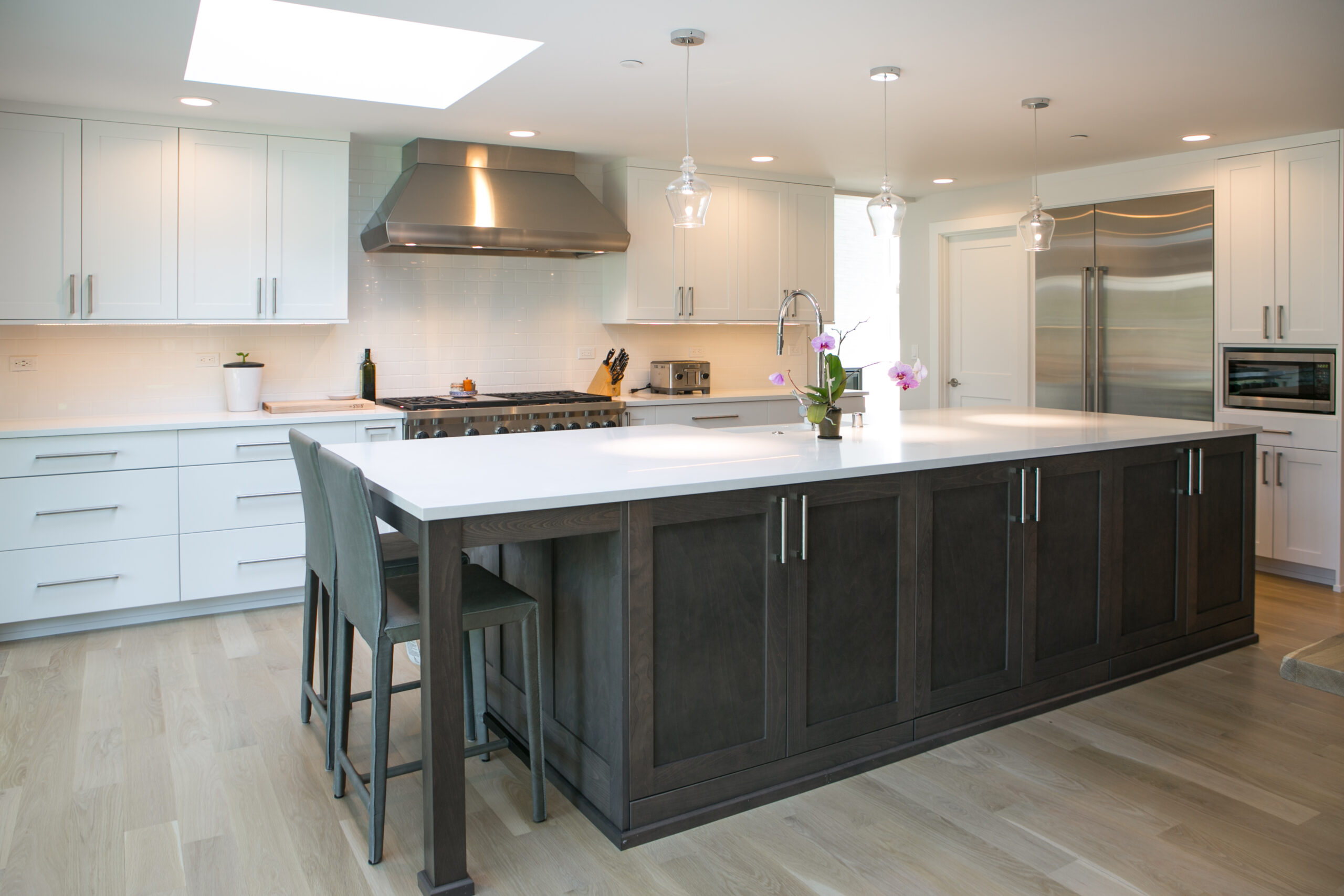 Modern kitchen with white cabinets, stainless steel appliances, a large dark wood island with a white countertop, two gray barstools, pendant lights, and light wood flooring.