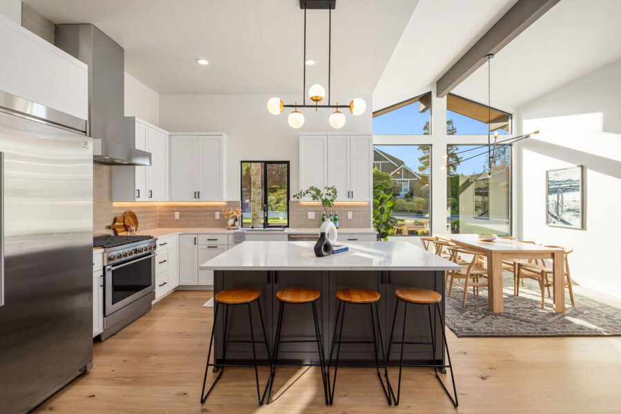 Modern kitchen with a large island, three wooden stools, stainless steel appliances, white cabinets, and pendant lighting. Sunlit dining area with wooden table and chairs is visible near large windows.