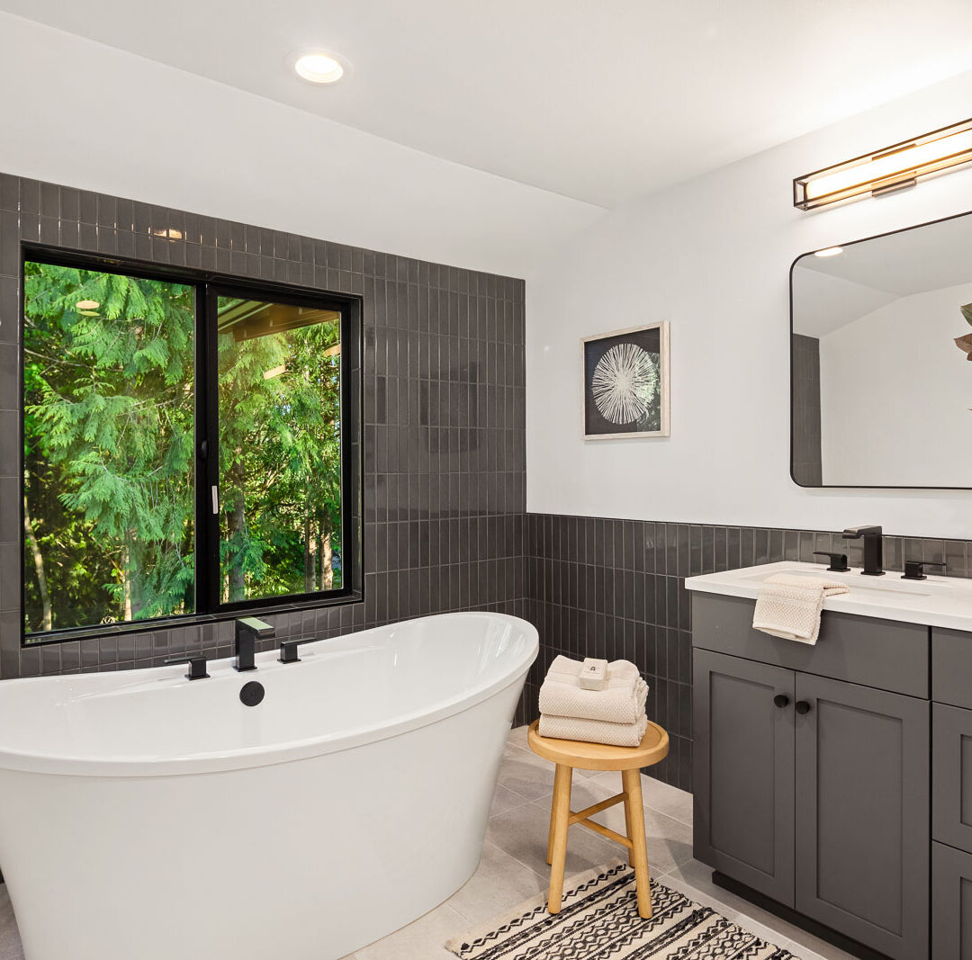 Modern bathroom with a freestanding white tub, dark gray tile wall, black-framed window showing greenery, gray vanity with a mirror, potted plant, towel on a stool, and patterned rug on a light floor.