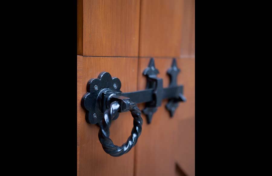 Close-up of a decorative black metal door latch with a twisted ring handle and ornate backplate, mounted on a wooden door with a warm brown finish.