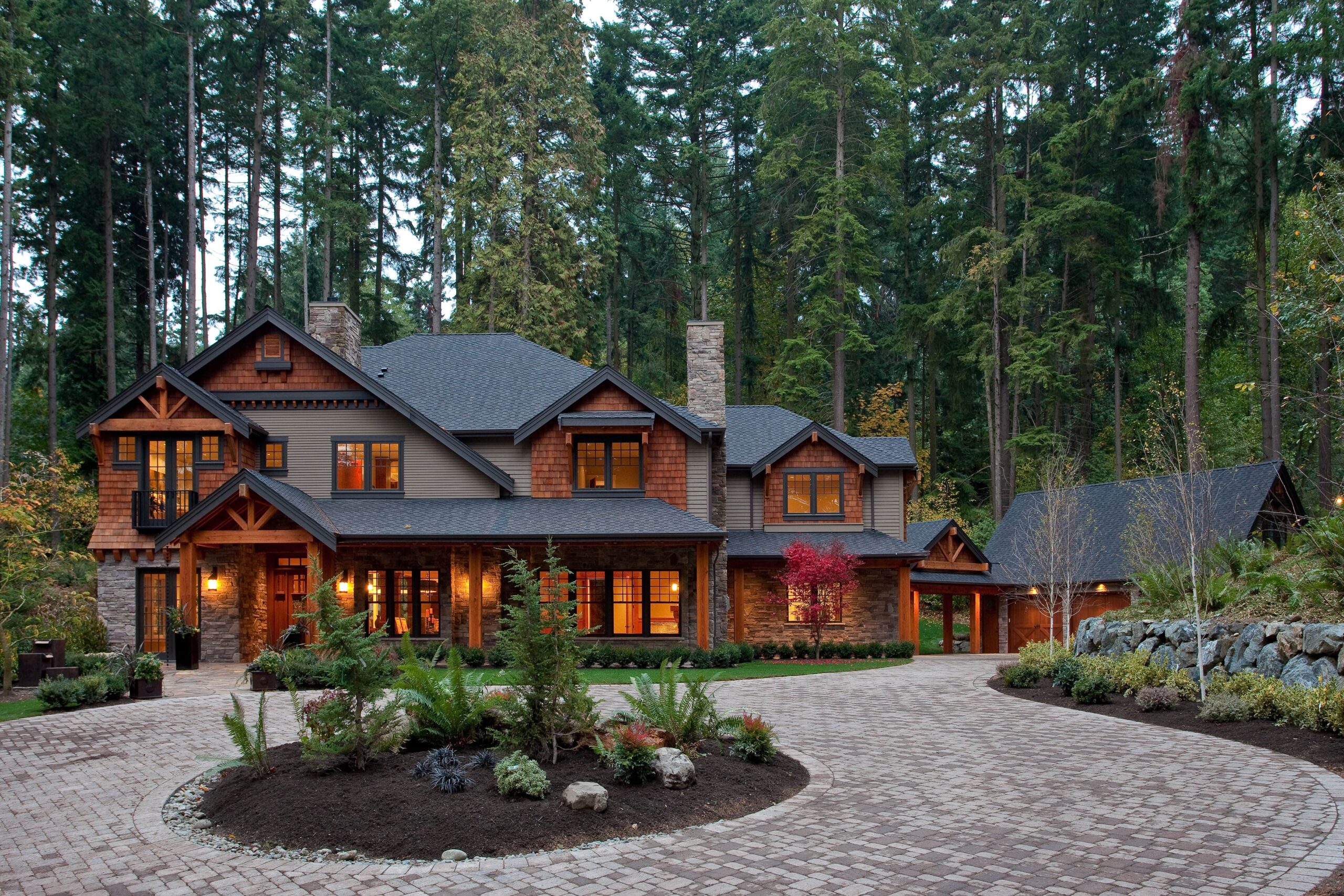 Large, rustic two-story house with wood and stone exterior, glowing warm lights inside, surrounded by tall trees, a stone chimney, landscaped yard, and a circular brick driveway in a forest setting.