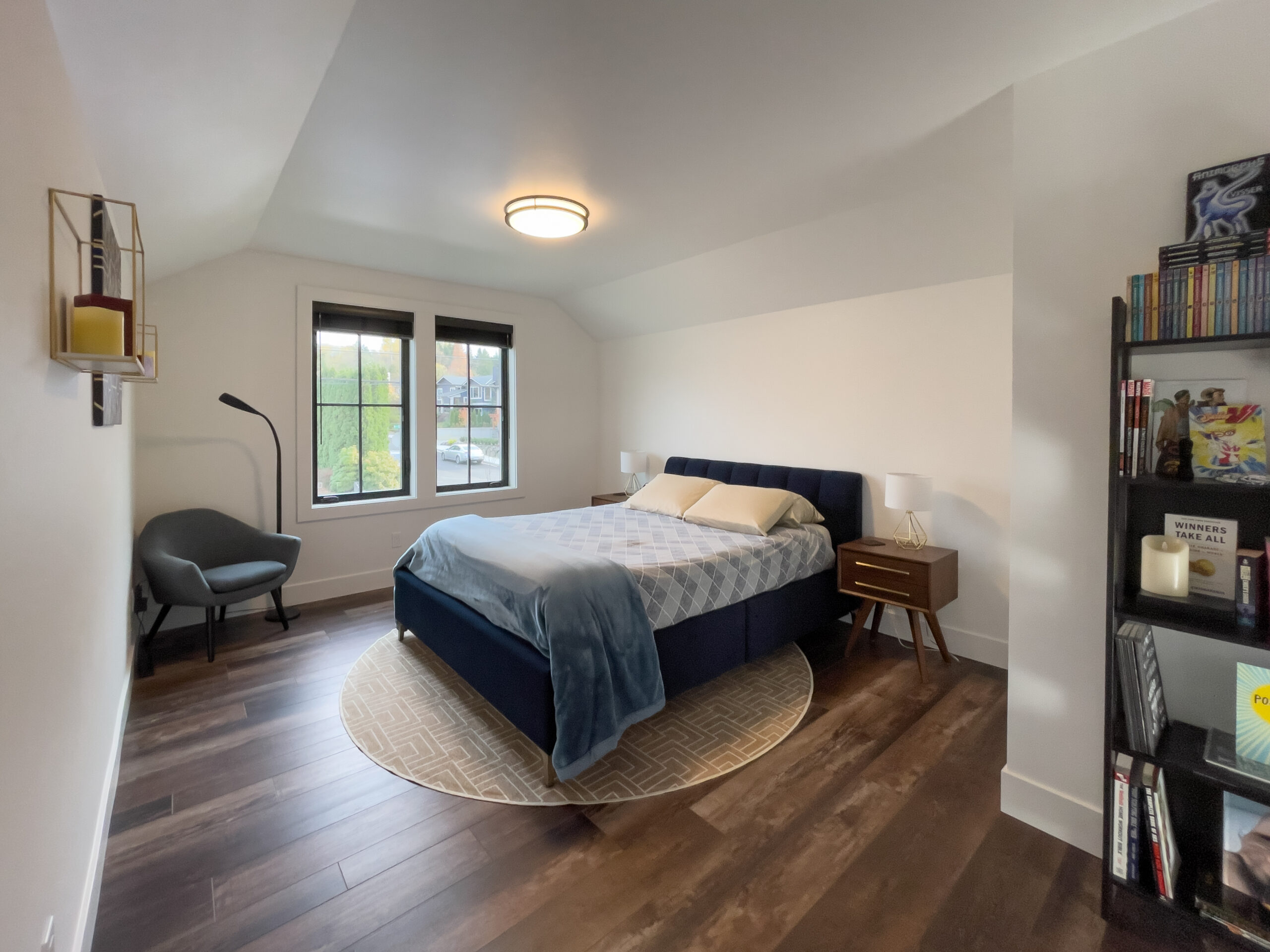A modern bedroom with a dark blue bed, two nightstands, and a round rug on wooden flooring. The room has a window with a view outside, a black armchair with a floor lamp, and a bookshelf on the right.
